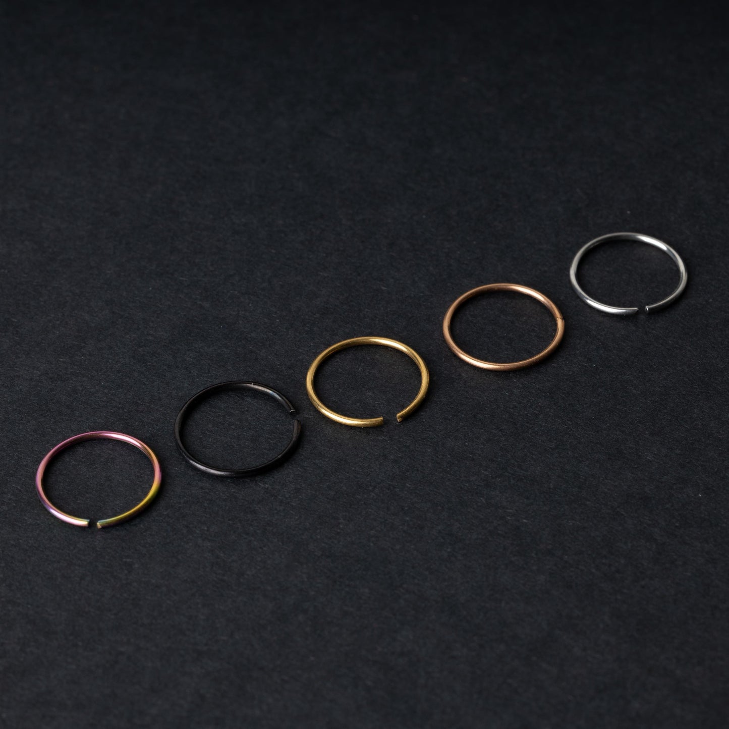 Five thin seamless hoops in rainbow, black, gold, rose gold, and silver finishes, displayed on a dark background.