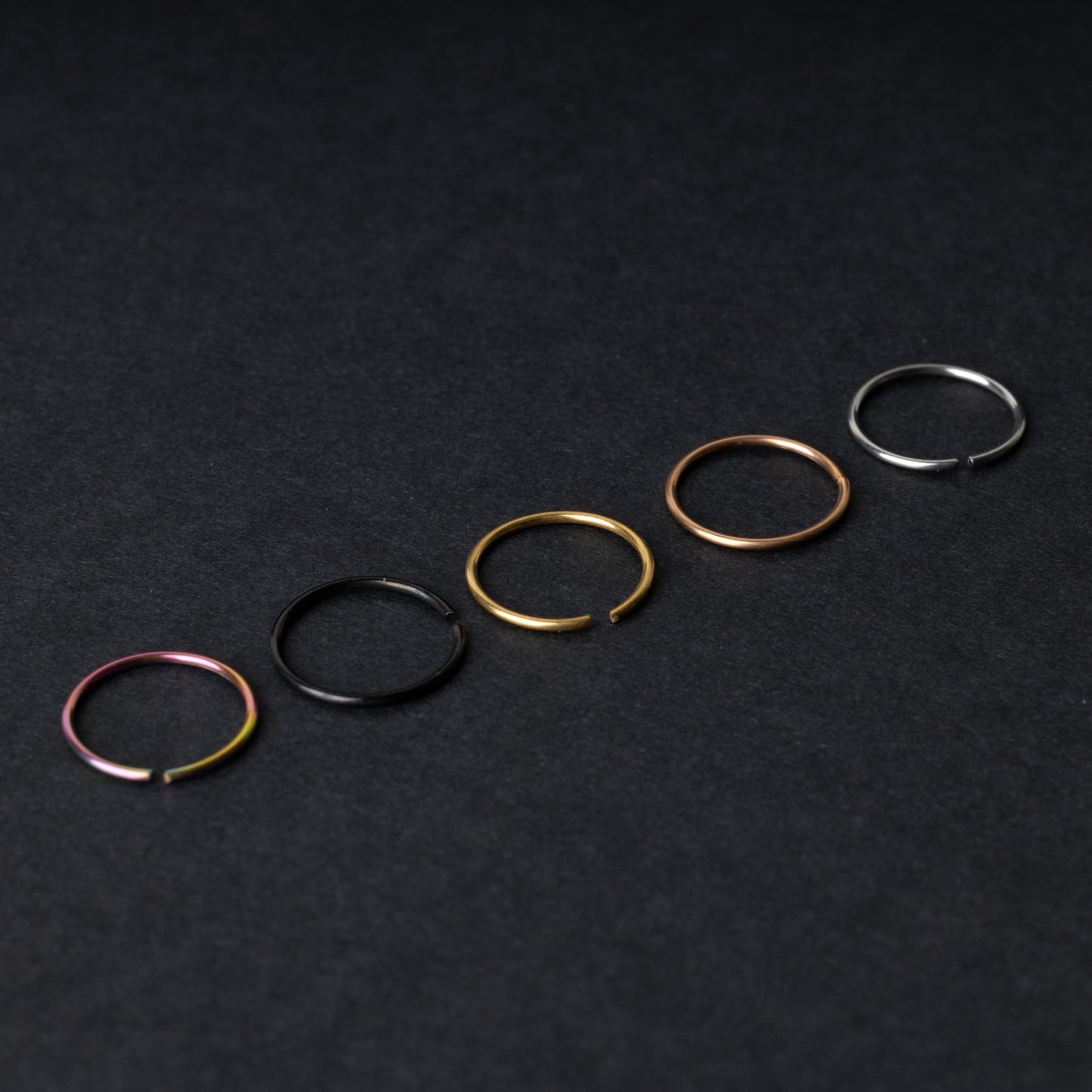 Five thin seamless hoops in rainbow, black, gold, rose gold, and silver finishes, displayed on a dark background.