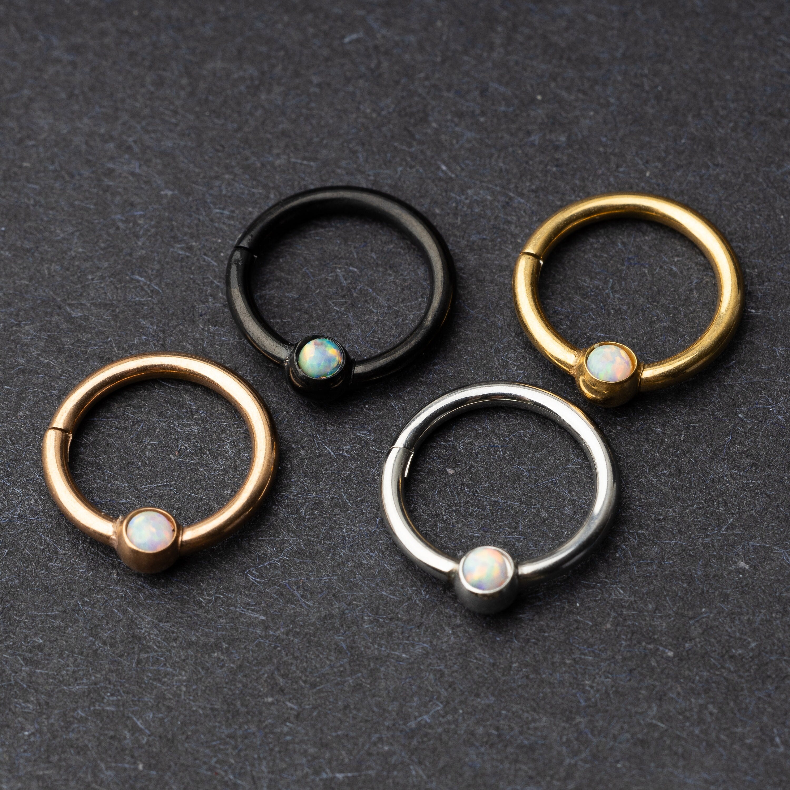 Four seamless rings in black, gold, rose gold, and silver finishes, each featuring an opalescent bead, displayed on a dark background.