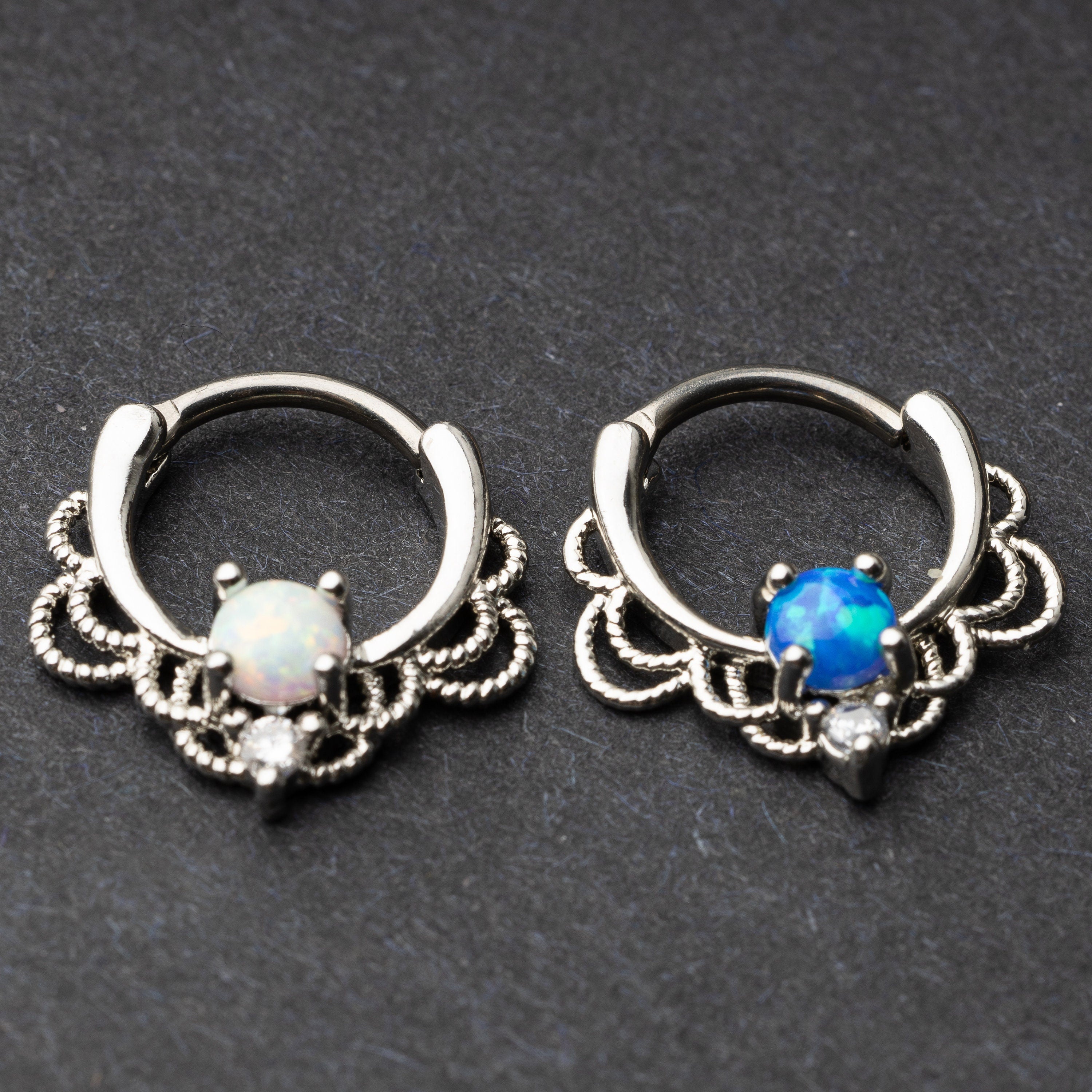 Two silver septum clickers with intricate lace designs. One features a white opal centerpiece, and the other showcases a vibrant blue stone, both with a clear cubic zirconia accent.