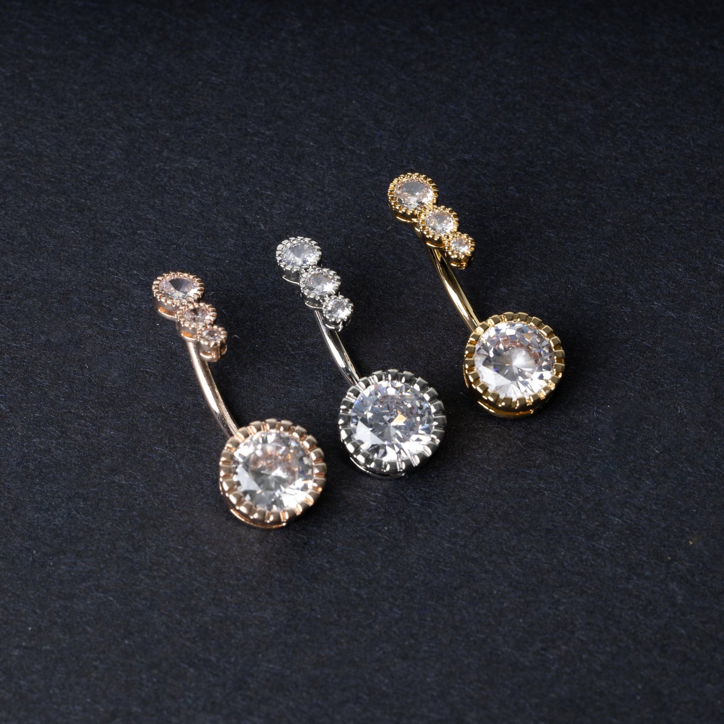 Double-jeweled belly rings in gold, silver, and rose gold finishes with large and small crystal accents, displayed on a dark background.
