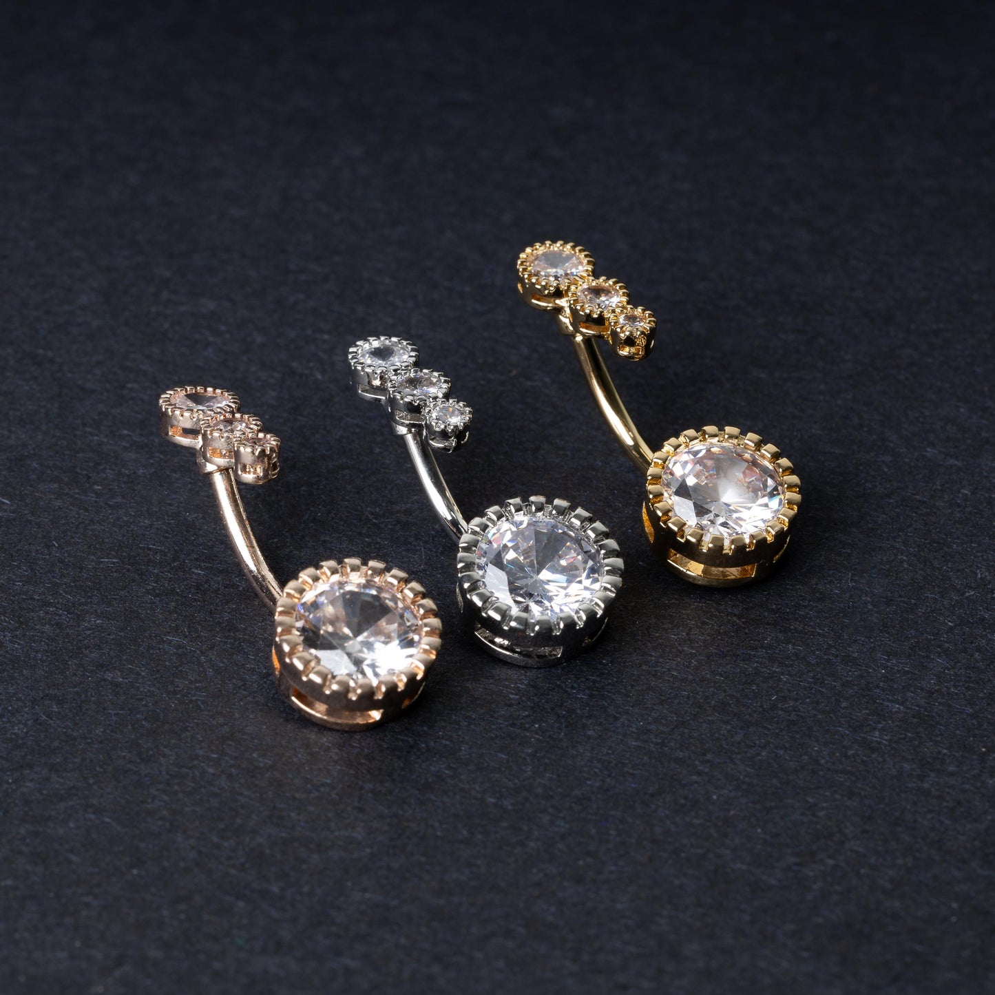 Double-jeweled belly rings in gold, silver, and rose gold finishes with large and small crystal accents, displayed on a dark background.