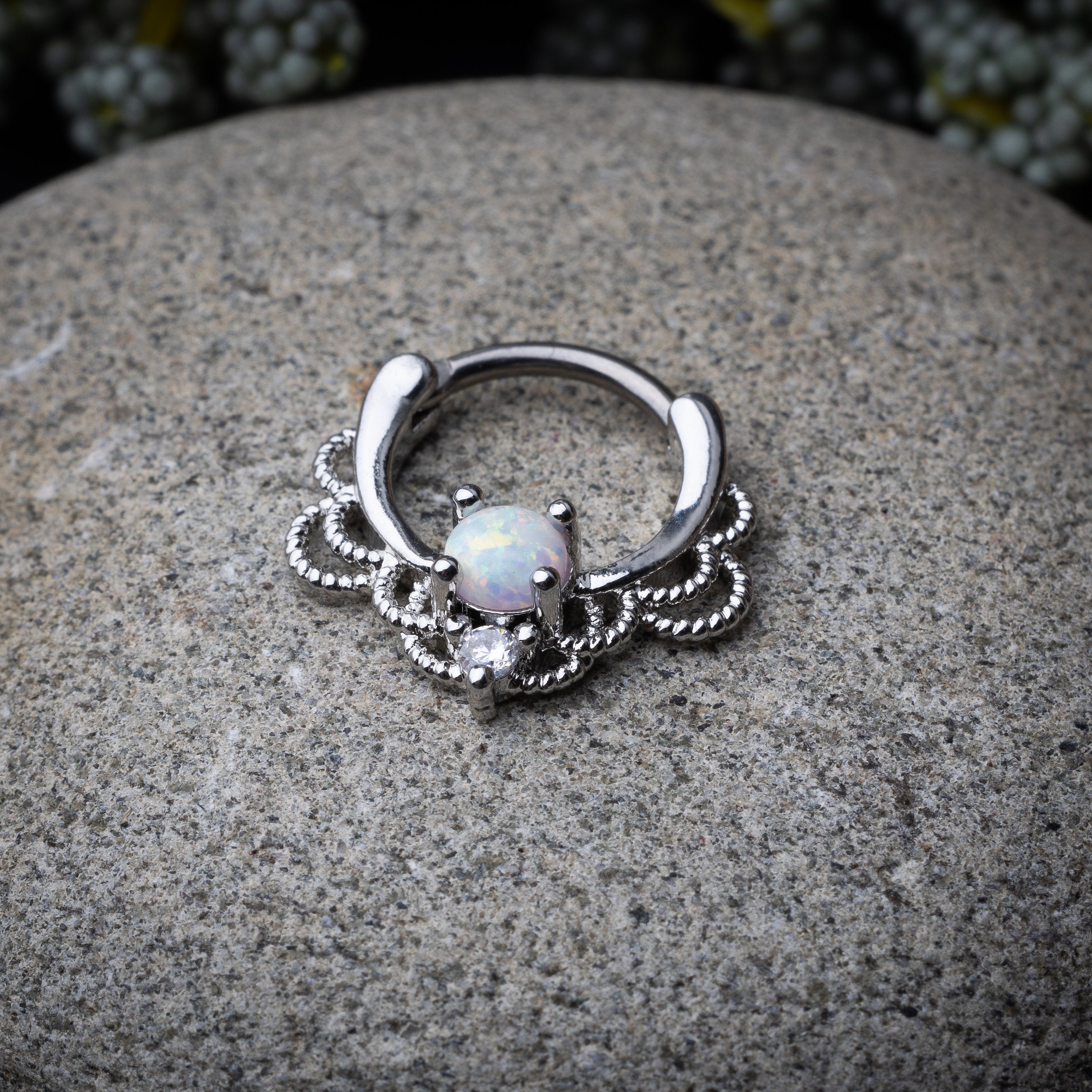 Silver septum clicker with intricate lace design displayed on a stone background.