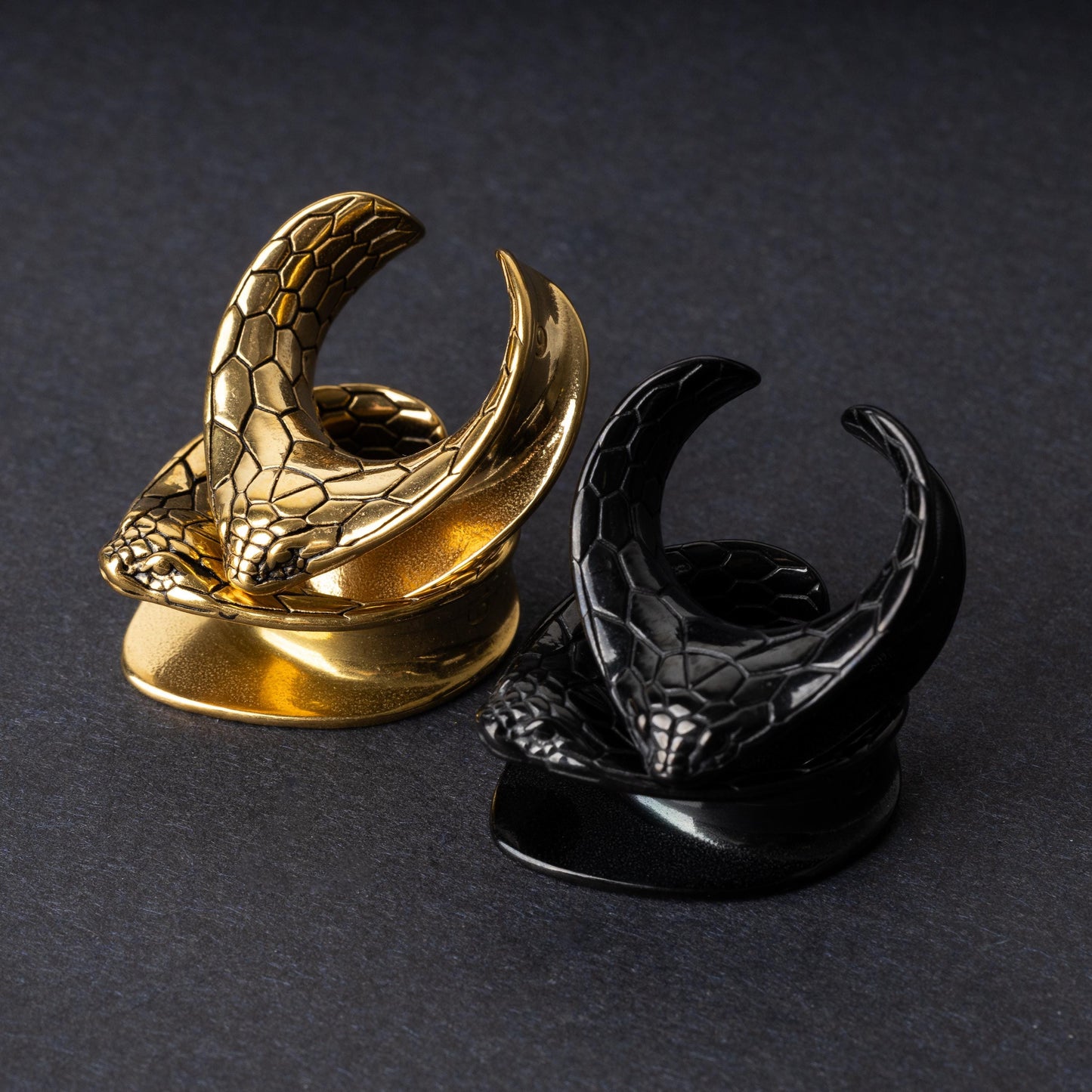 A pair of snakehead saddle tunnels for stretched ears in black and gold finishes, displayed on a dark background.