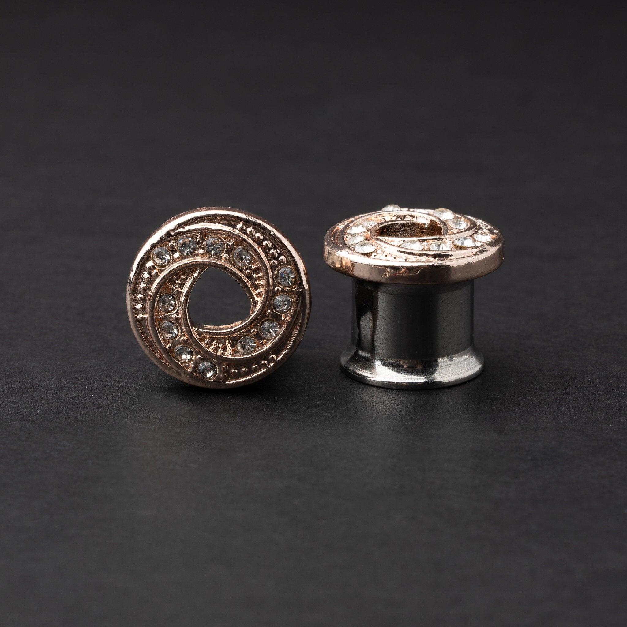 A pair of rose gold-tone stainless steel screw-fit tunnels with an infinity swirl design encrusted with clear crystals, displayed against a dark background.