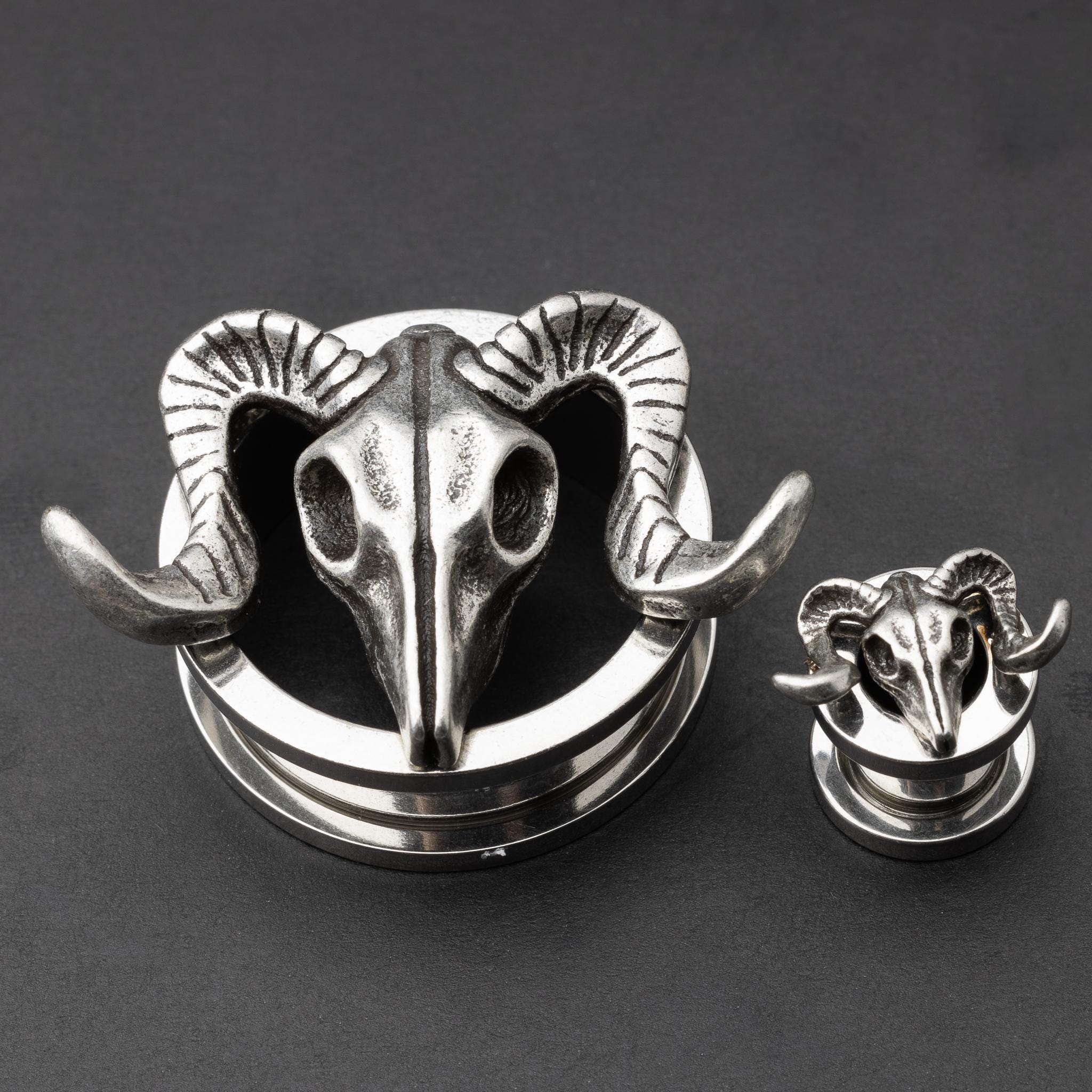 A pair of stainless steel saddle plugs featuring a 3D ram skull design, displayed on a dark, textured background.