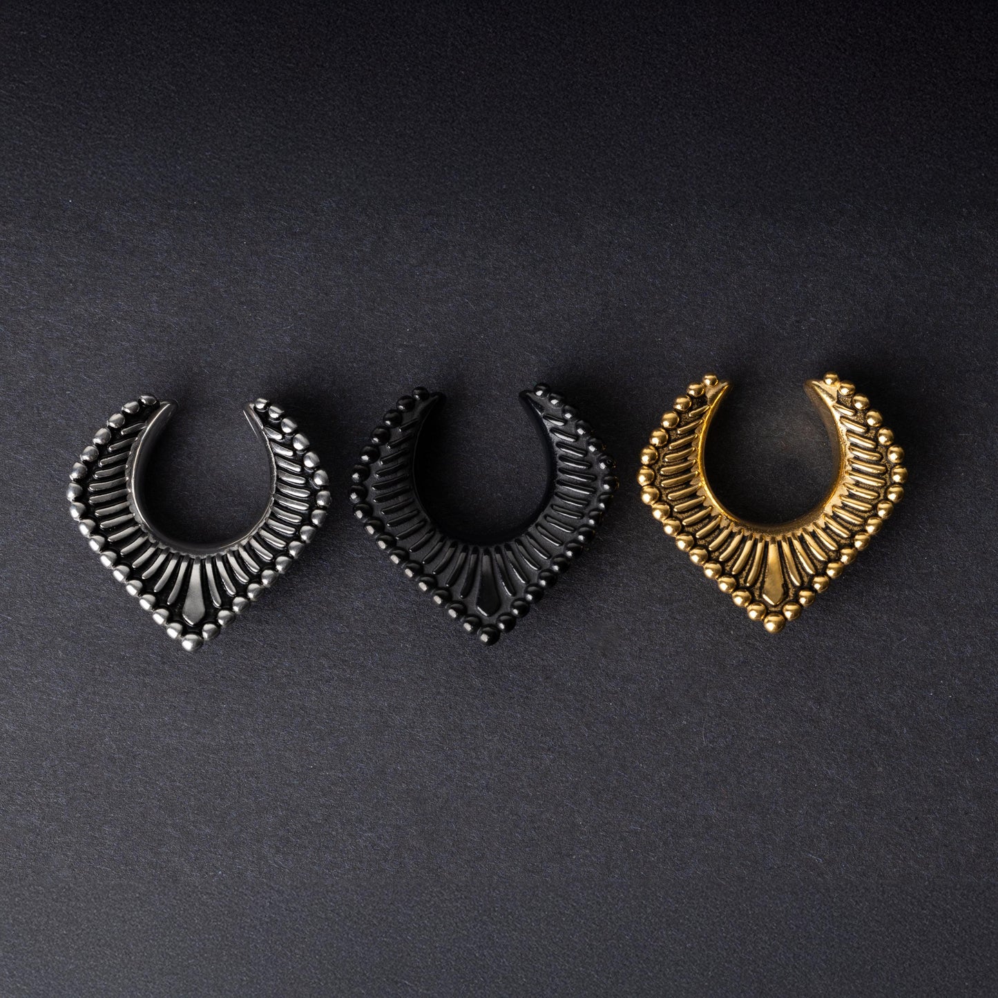 Three V-shaped stainless steel clickers in silver, black, and gold finishes arranged on a dark textured background.