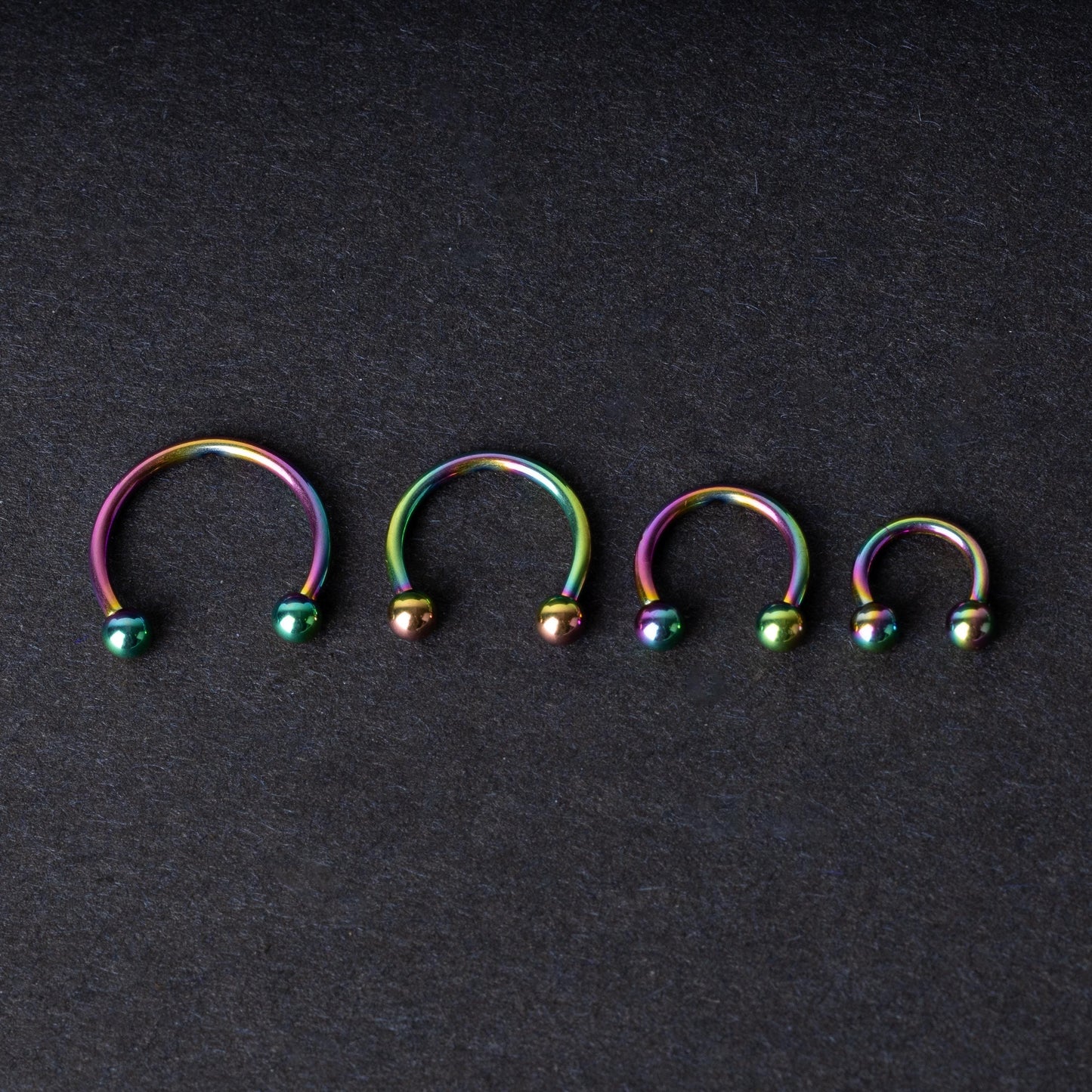 Close-up of four anodized rainbow horseshoe rings in varying sizes, with vibrant, multicolor finishes, displayed on a dark background.