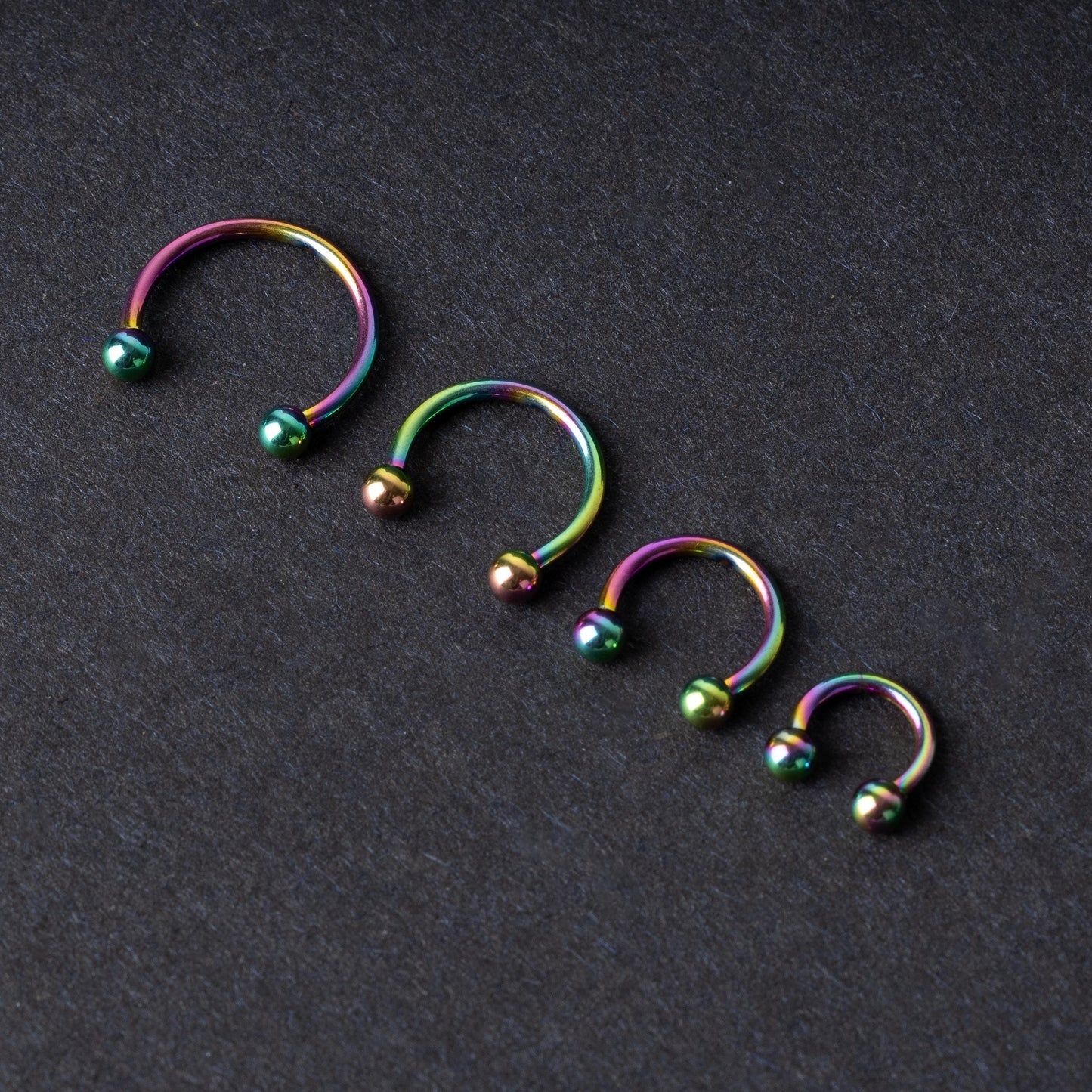 Close-up of four anodized rainbow horseshoe rings in varying sizes, with vibrant, multicolor finishes, displayed on a dark background.