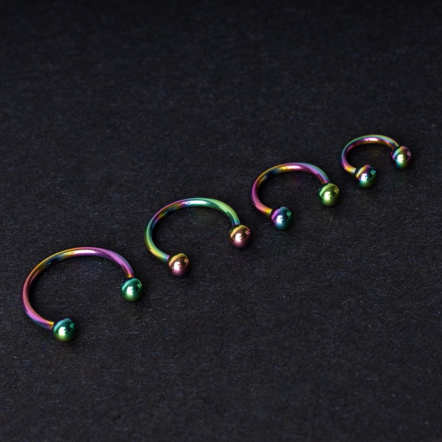 Close-up of four anodized rainbow horseshoe rings in varying sizes, with vibrant, multicolor finishes, displayed on a dark background.