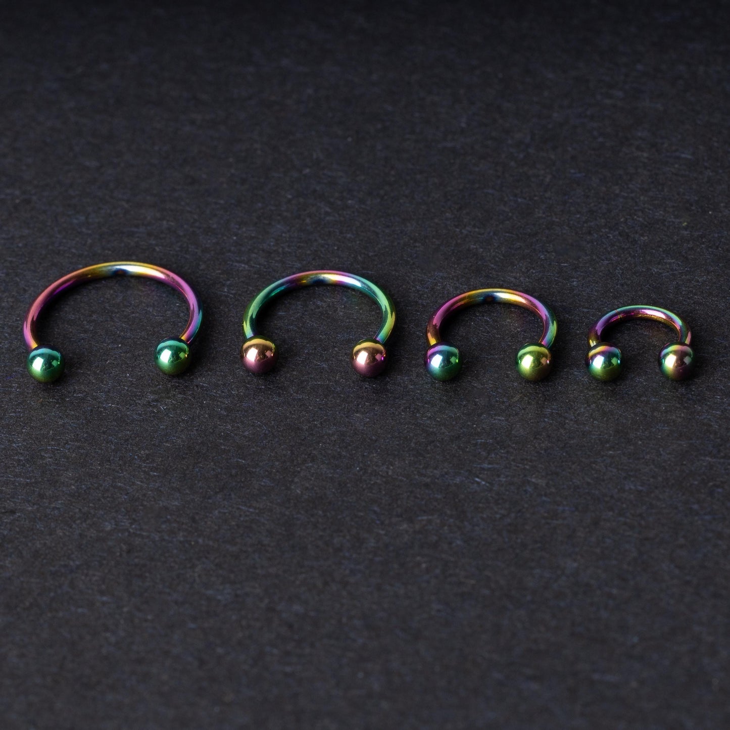 Close-up of four anodized rainbow horseshoe rings in varying sizes, with vibrant, multicolor finishes, displayed on a dark background.