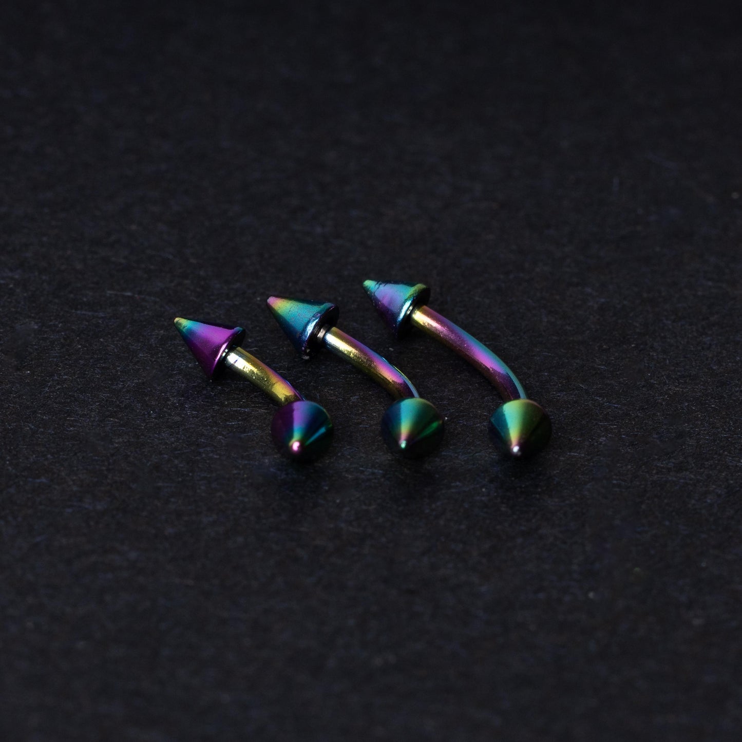 Three rainbow titanium curved barbells with cone-shaped ends, displayed on a dark textured background, showcasing their vivid color gradient and sleek design.