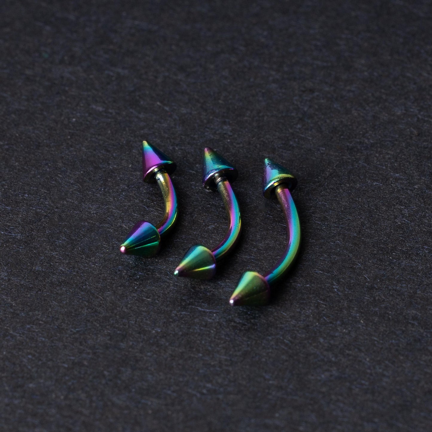 Three rainbow titanium curved barbells with cone-shaped ends, displayed on a dark textured background, showcasing their vivid color gradient and sleek design.