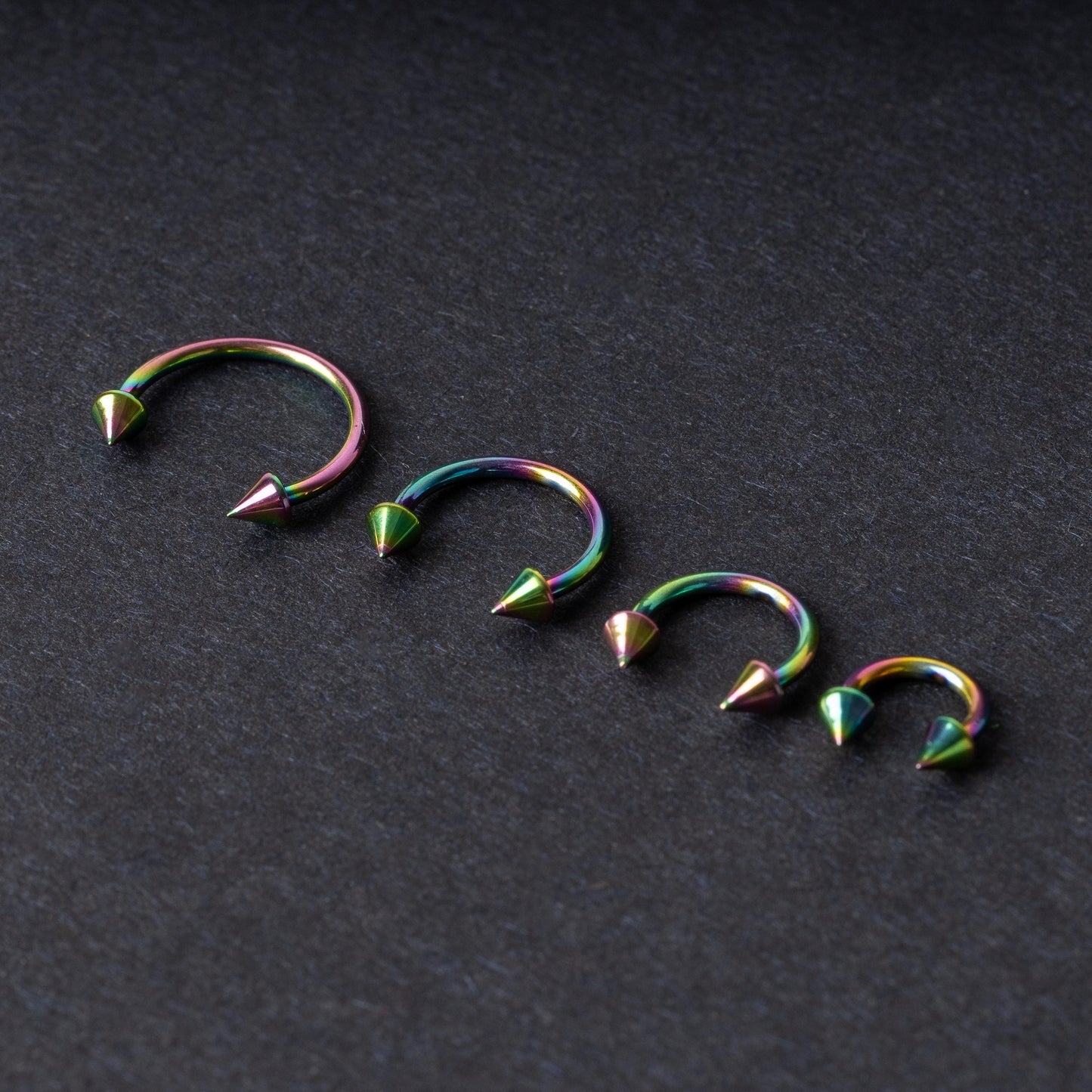 Titanium horseshoe hoops with spike ends, featuring a colorful rainbow finish, displayed on a dark textured background.