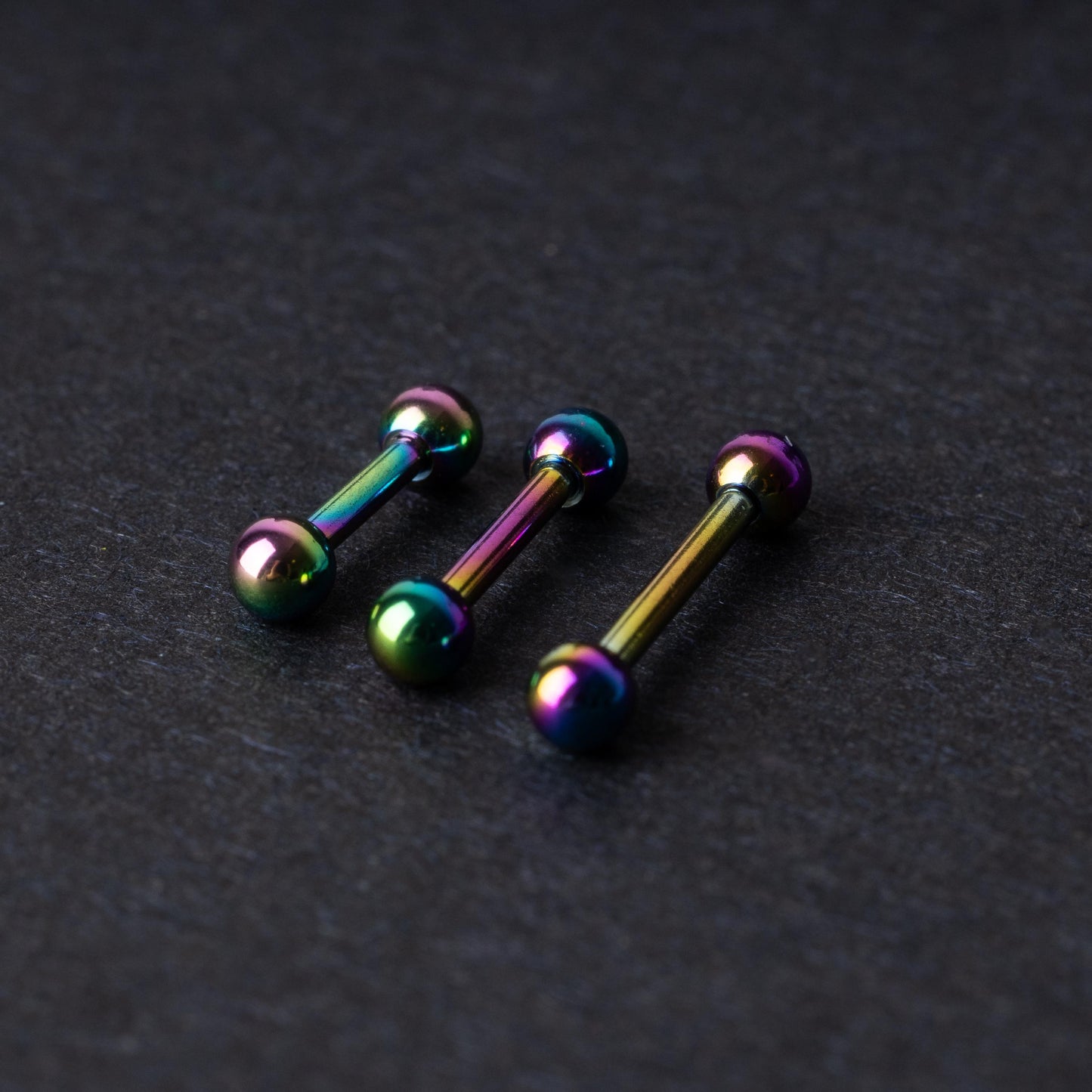 Rainbow stainless steel barbells with an anodized finish on a dark background.