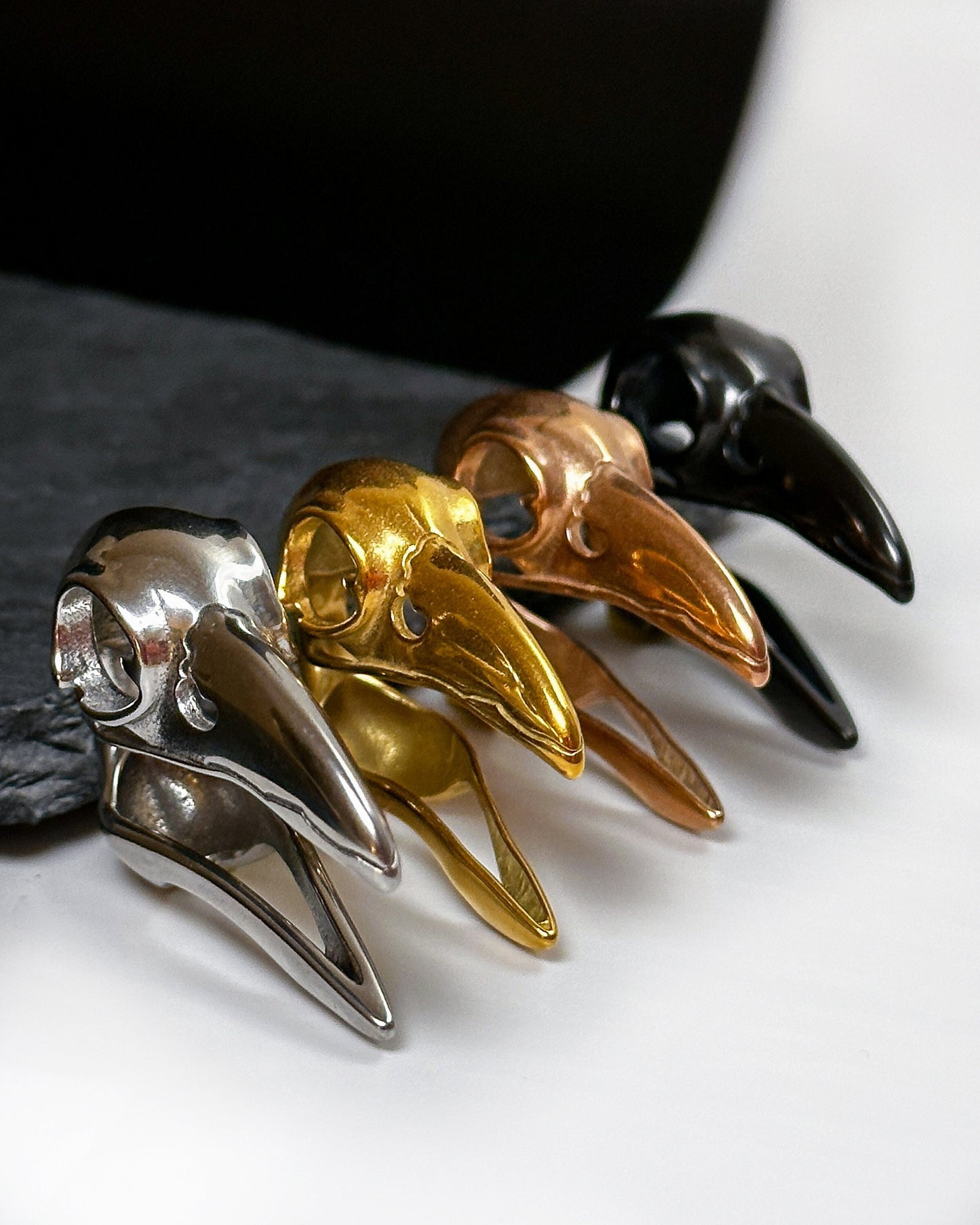 Four raven skull saddle plugs in black, silver, gold, and rose gold finishes, elegantly displayed on a dark background.