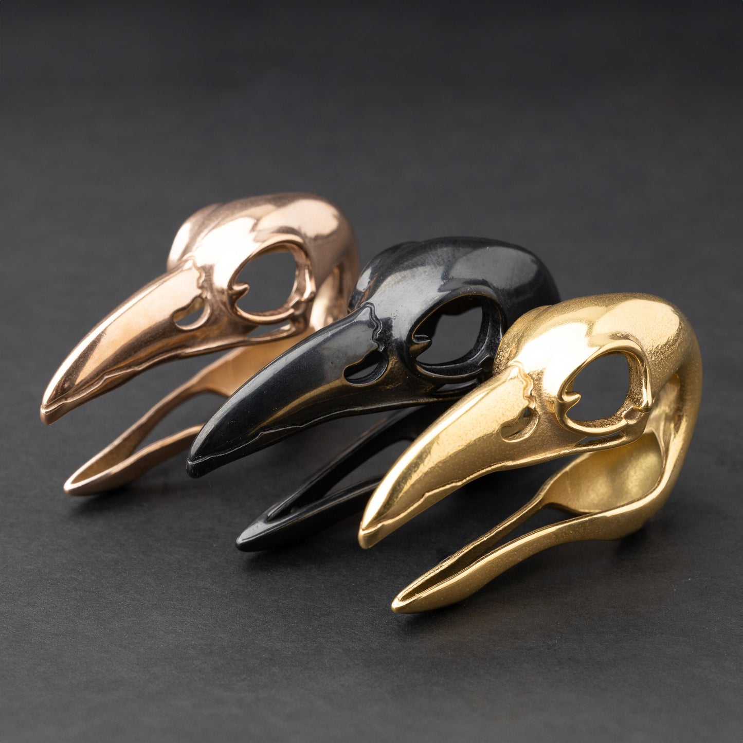 Three raven skull saddle plugs in black, gold, and rose gold finishes, elegantly displayed on a dark background.