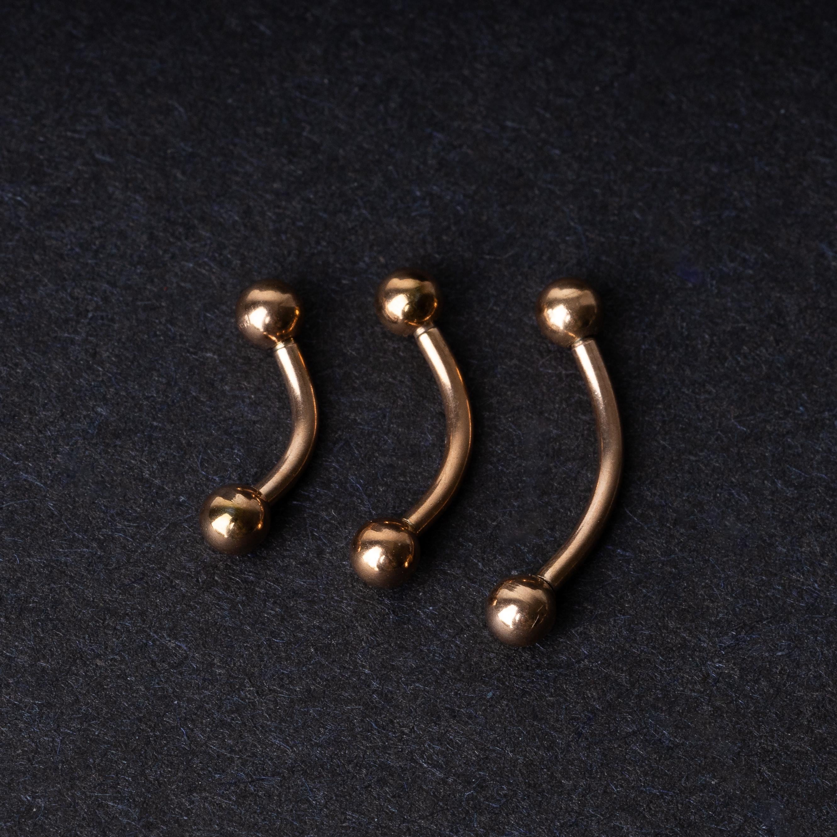 Three rose gold-tone stainless steel curved barbells of varying sizes displayed on a dark background.