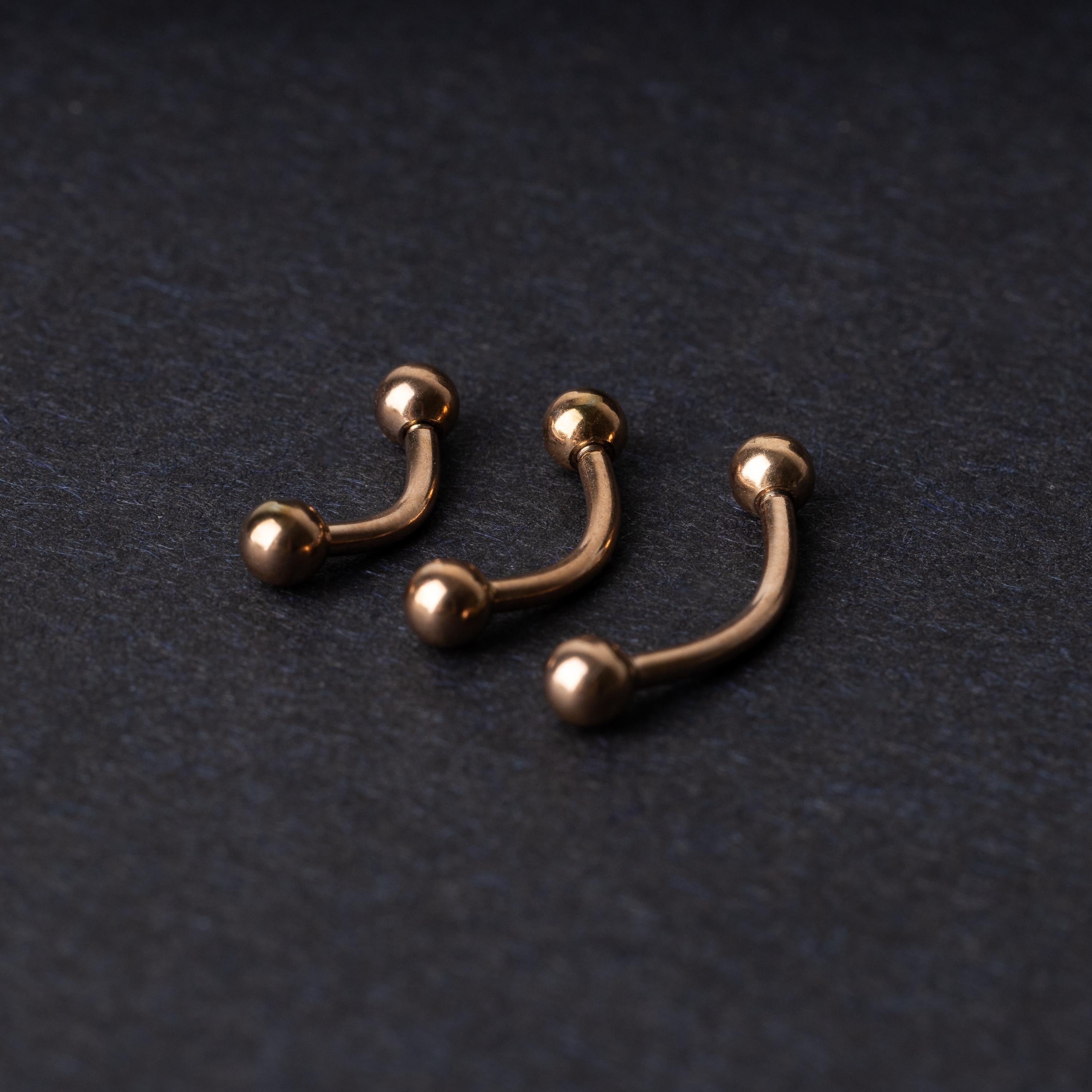Three rose gold-tone stainless steel curved barbells of varying sizes displayed on a dark background.