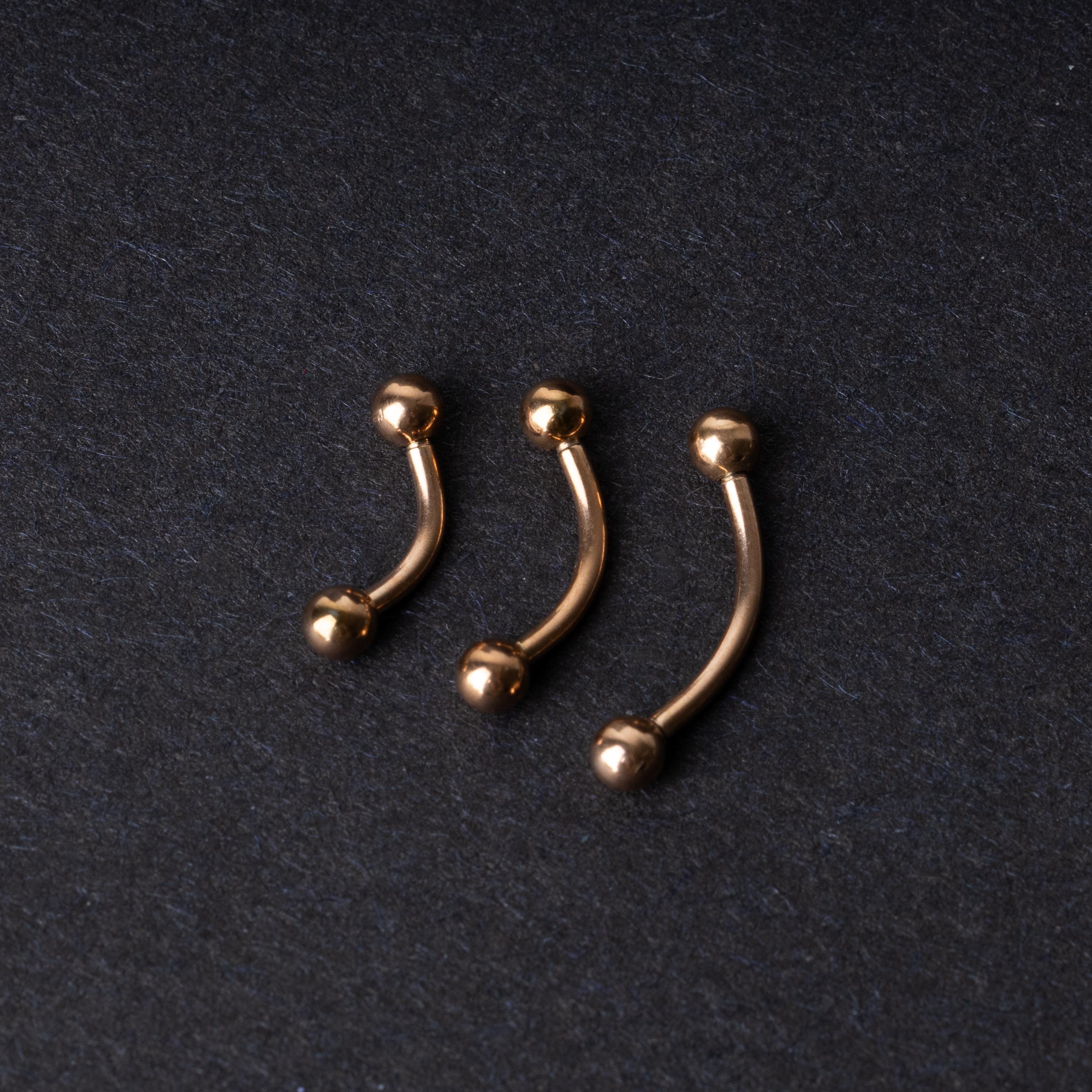 Three rose gold-tone stainless steel curved barbells of varying sizes displayed on a dark background.