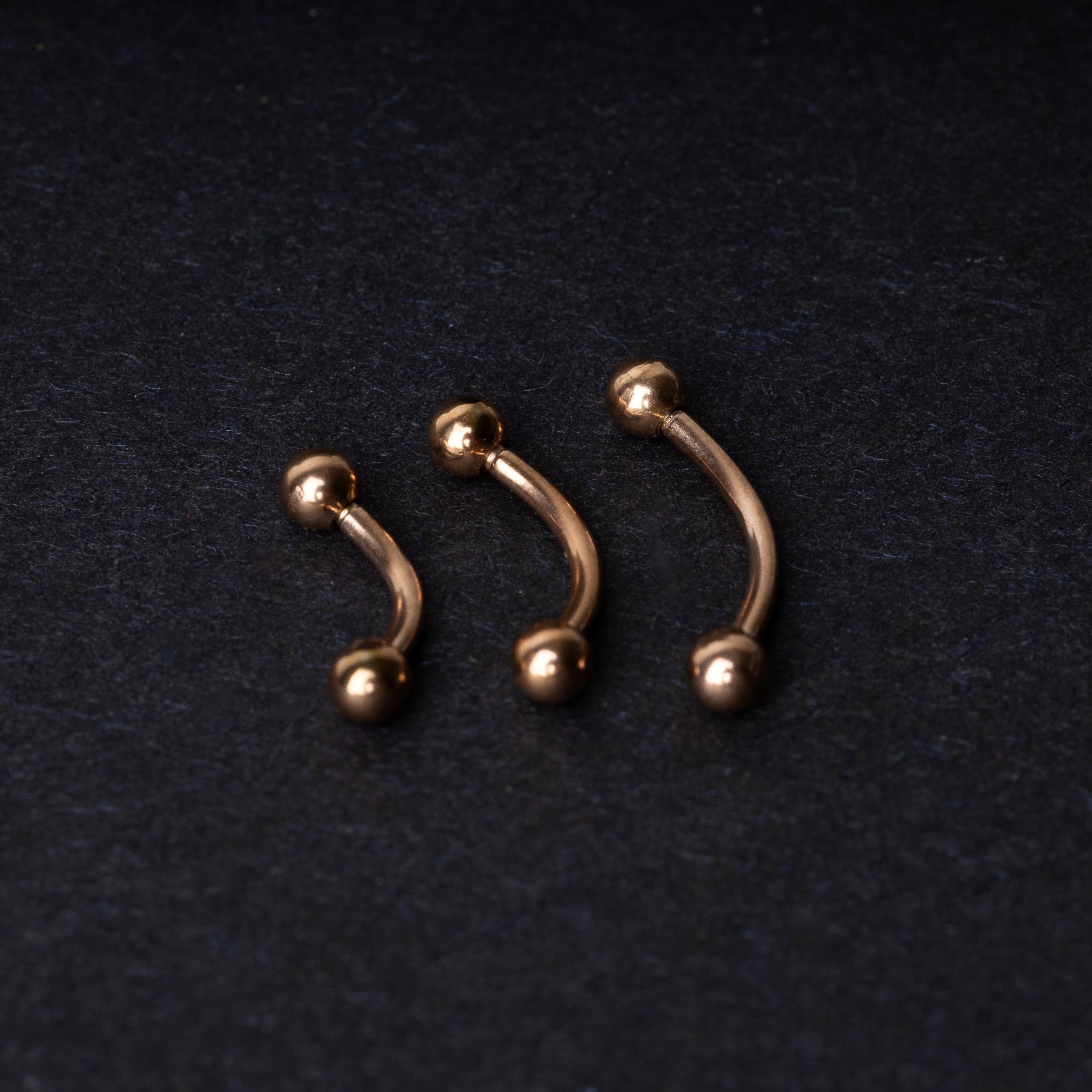 Three rose gold-tone stainless steel curved barbells of varying sizes displayed on a dark background.