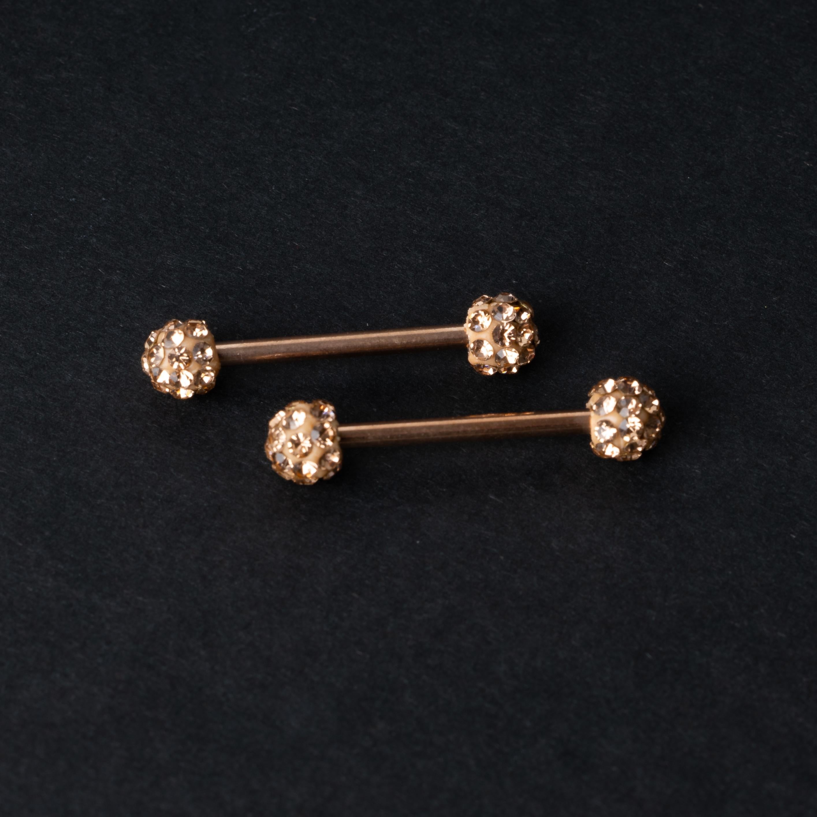 Barbells with textured crystal-encrusted ends in rose gold displayed on a dark background.