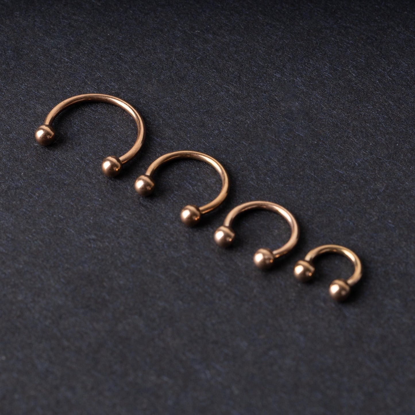 Rose Gold-tone horseshoe rings displayed in a row on a dark background, showcasing various sizes for septum or body piercings.