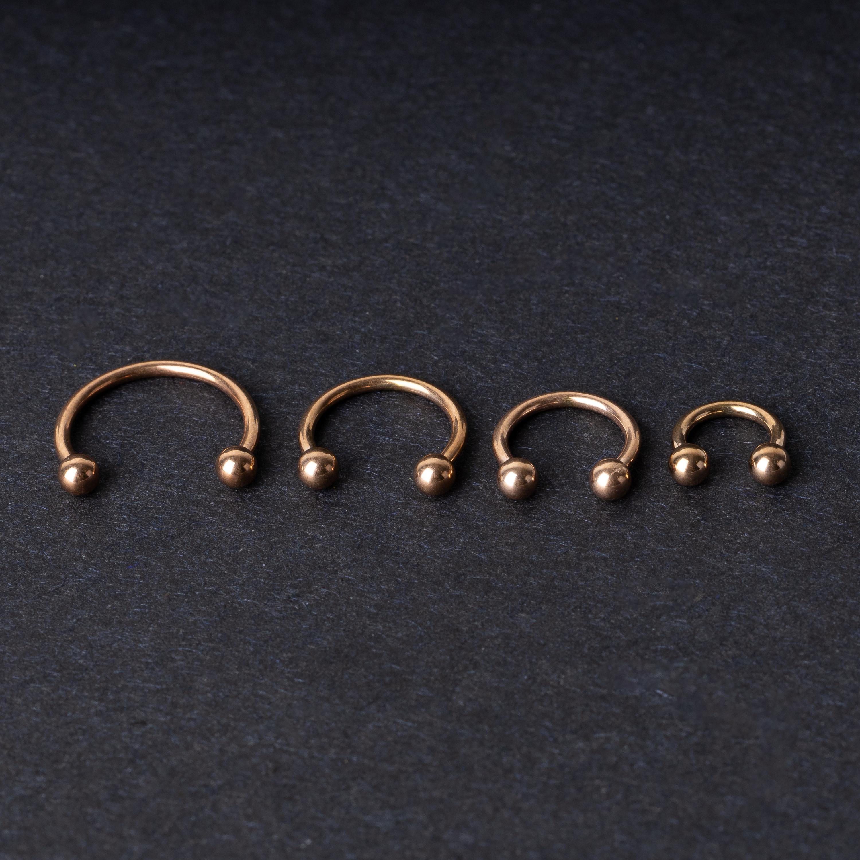 Rose Gold-tone horseshoe rings displayed in a row on a dark background, showcasing various sizes for septum or body piercings.