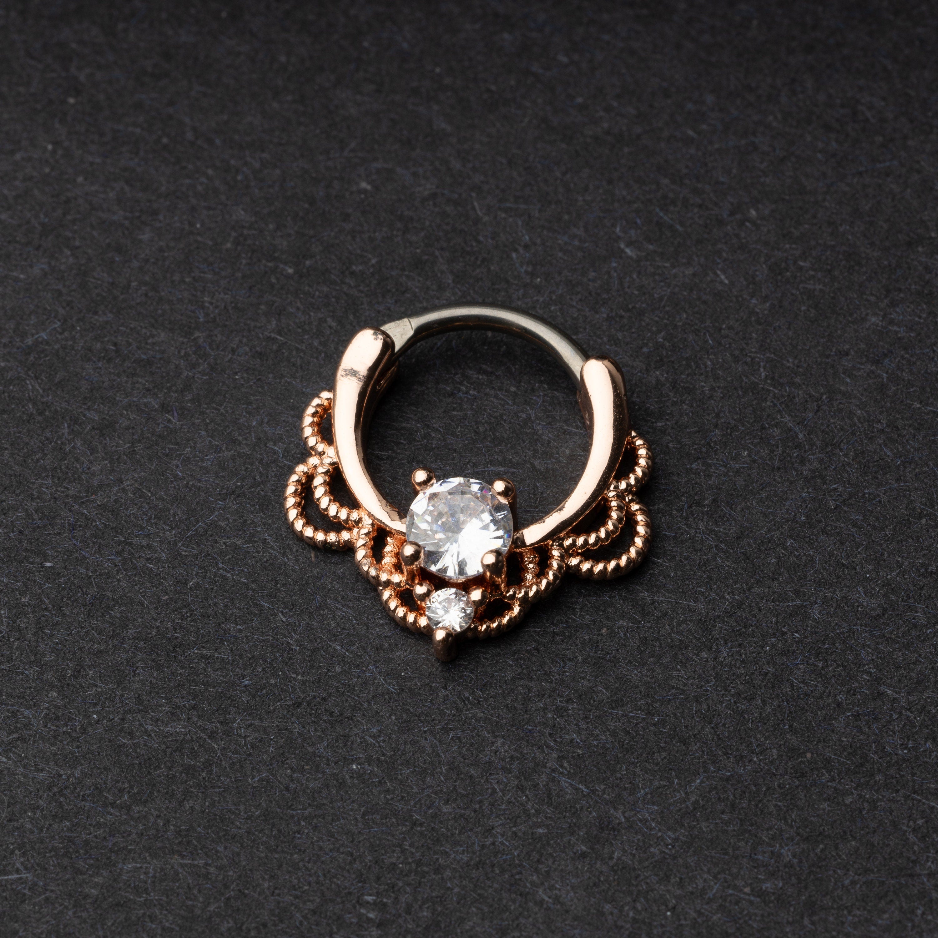 Rose gold ornate septum ring with a clear gem on a dark background.