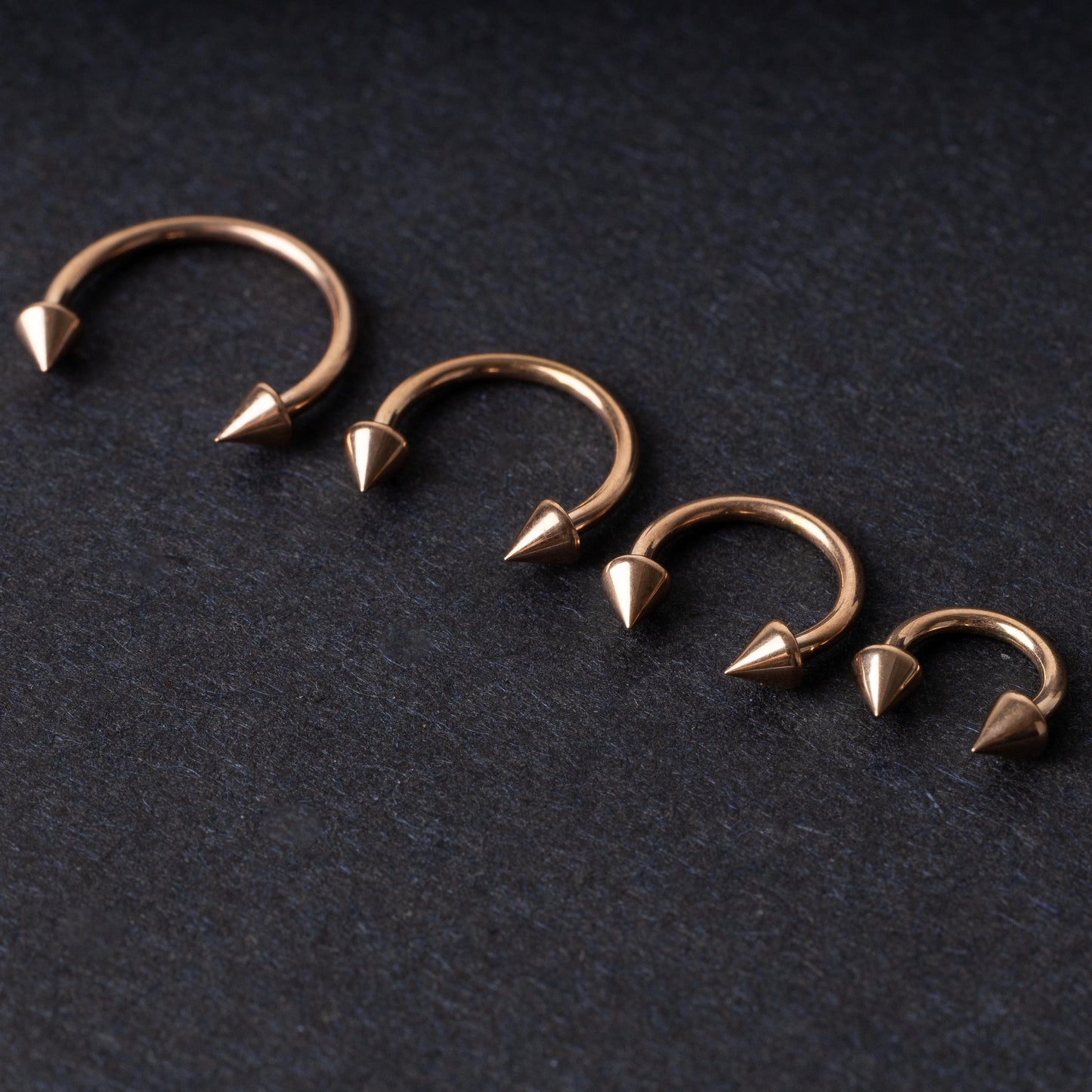 Four rose gold-tone horseshoe hoops with spike ends, arranged in descending size order on a dark textured background.
