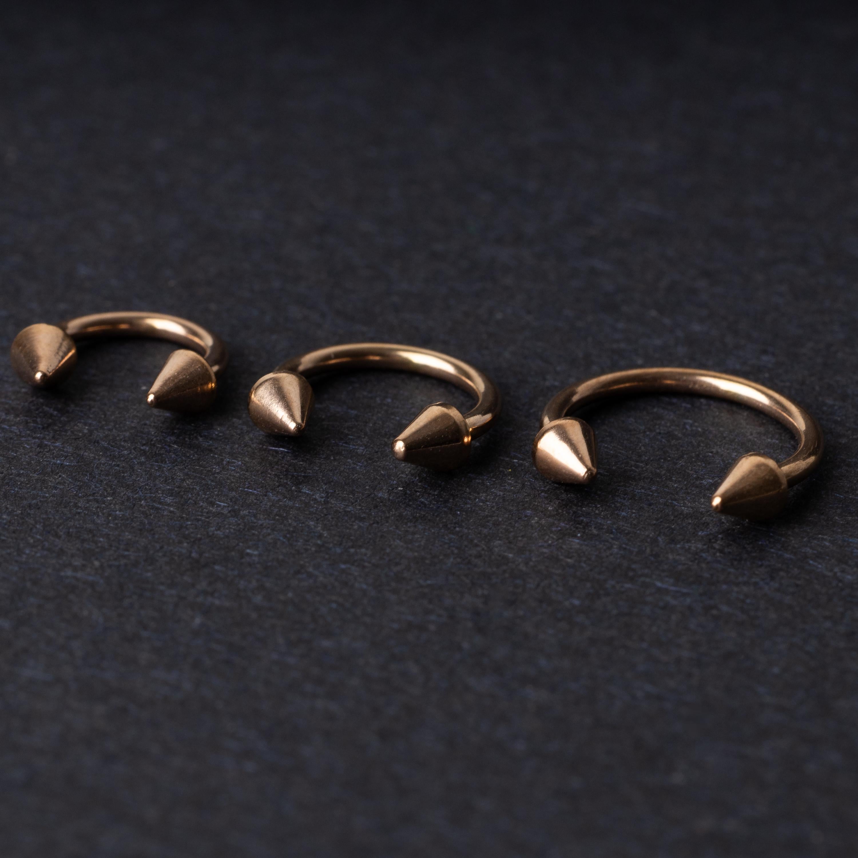 Three rose gold-tone horseshoe hoops with threaded spike ends arranged on a dark textured background.