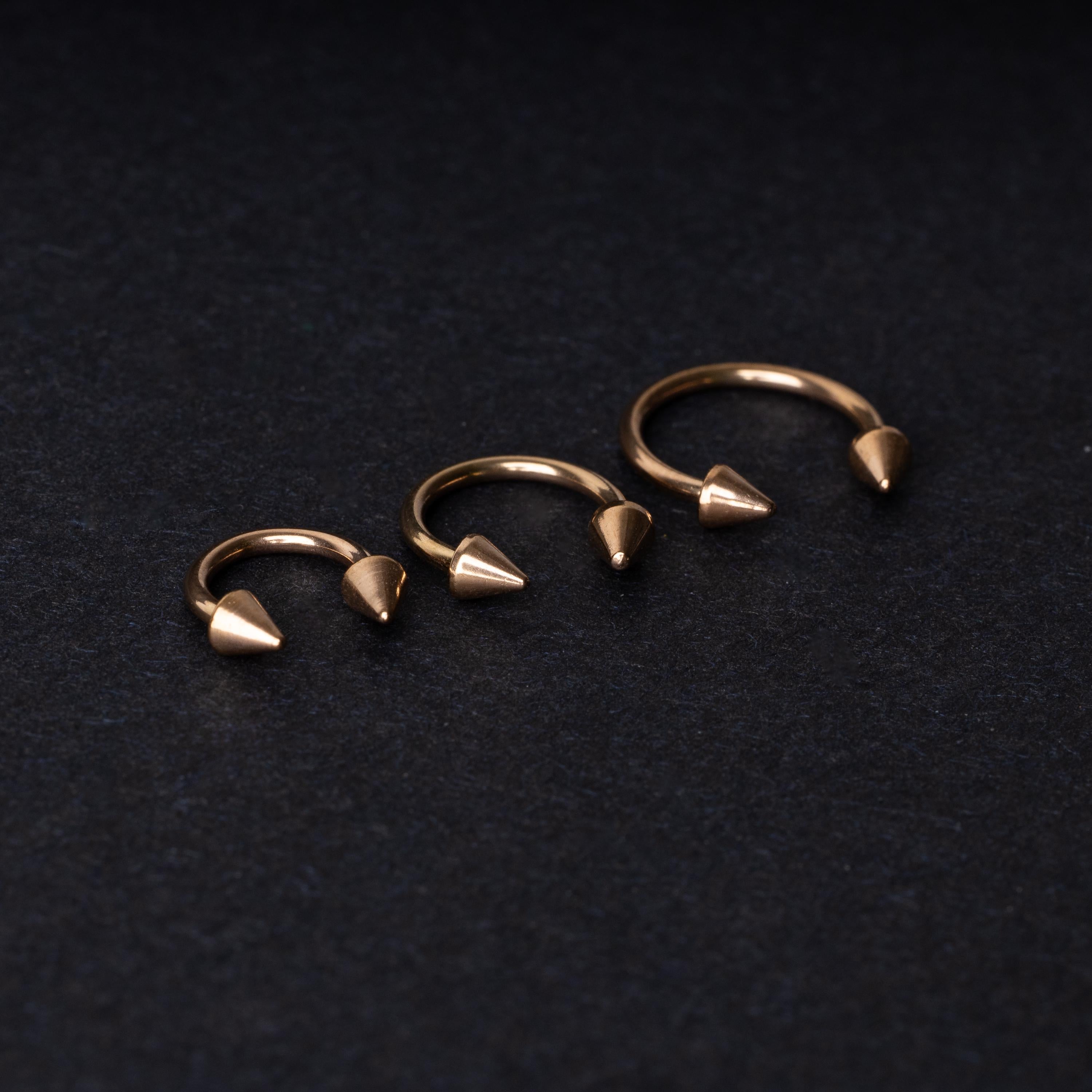 Three rose gold-tone horseshoe hoops with threaded spike ends arranged on a dark textured background.