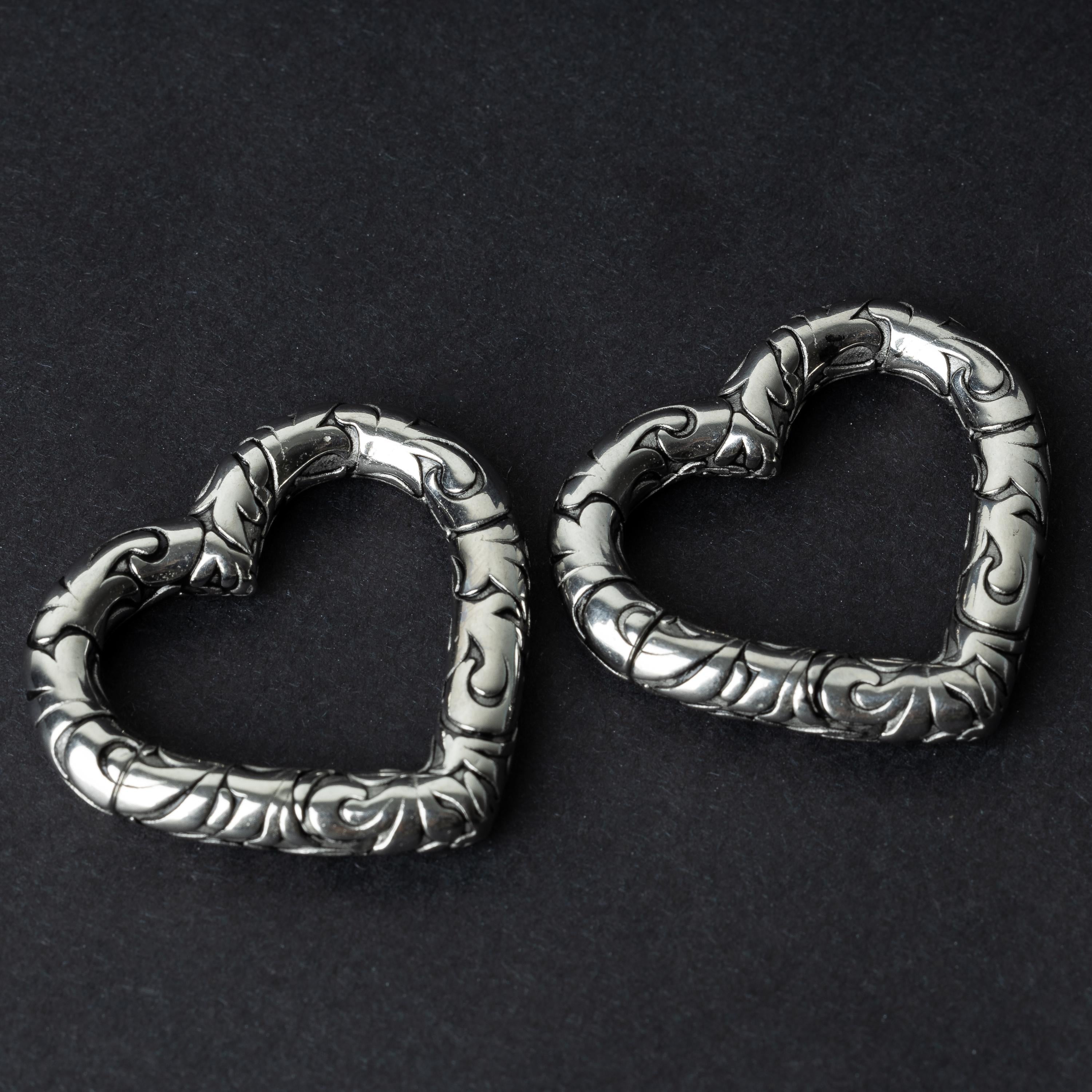 Two heart-shaped ear weights in silver, featuring intricate detailing, displayed on a dark background.