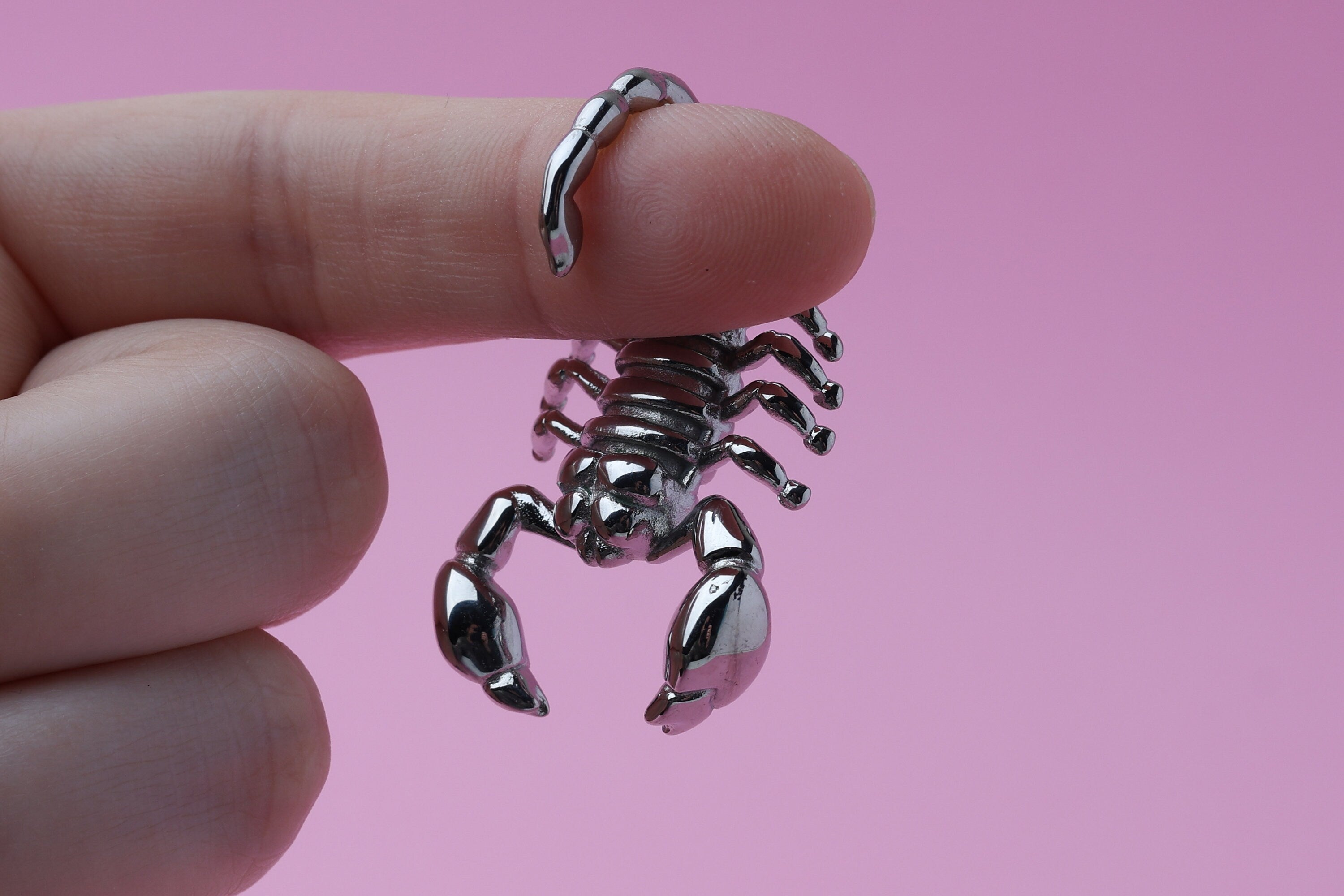 Scorpion-shaped ear hanger in silver and hung from a finger in front of a pink background, highlighting their detailed and edgy design.