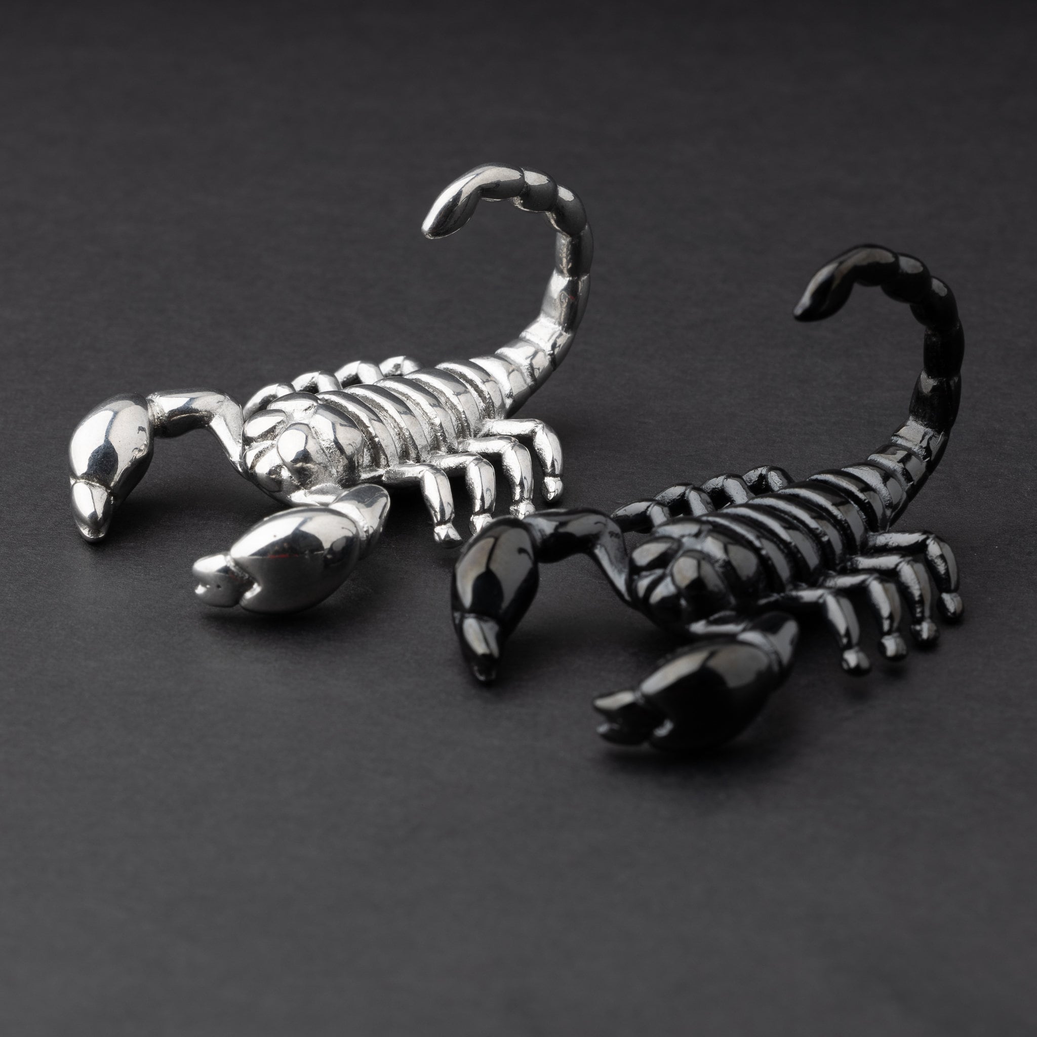 Two scorpion-shaped ear hangers, one in silver and one in black, displayed on a dark matte background, highlighting their detailed and edgy design.