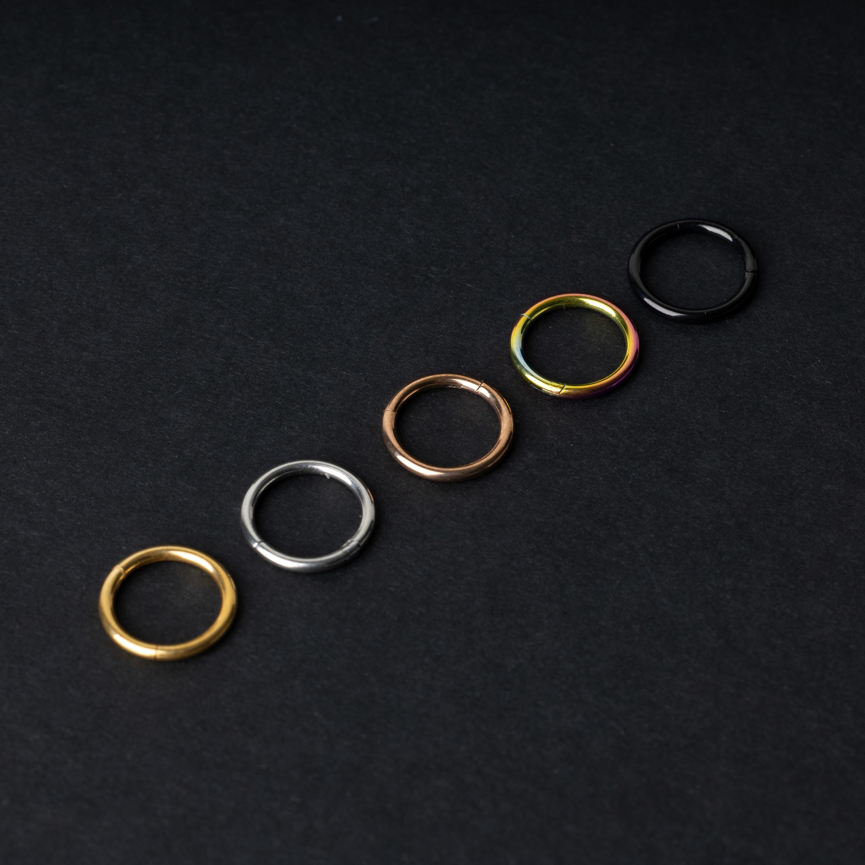 A selection of seamless clicker rings in various finishes – gold, silver, rose gold, rainbow, and black – displayed in a line on a dark background.