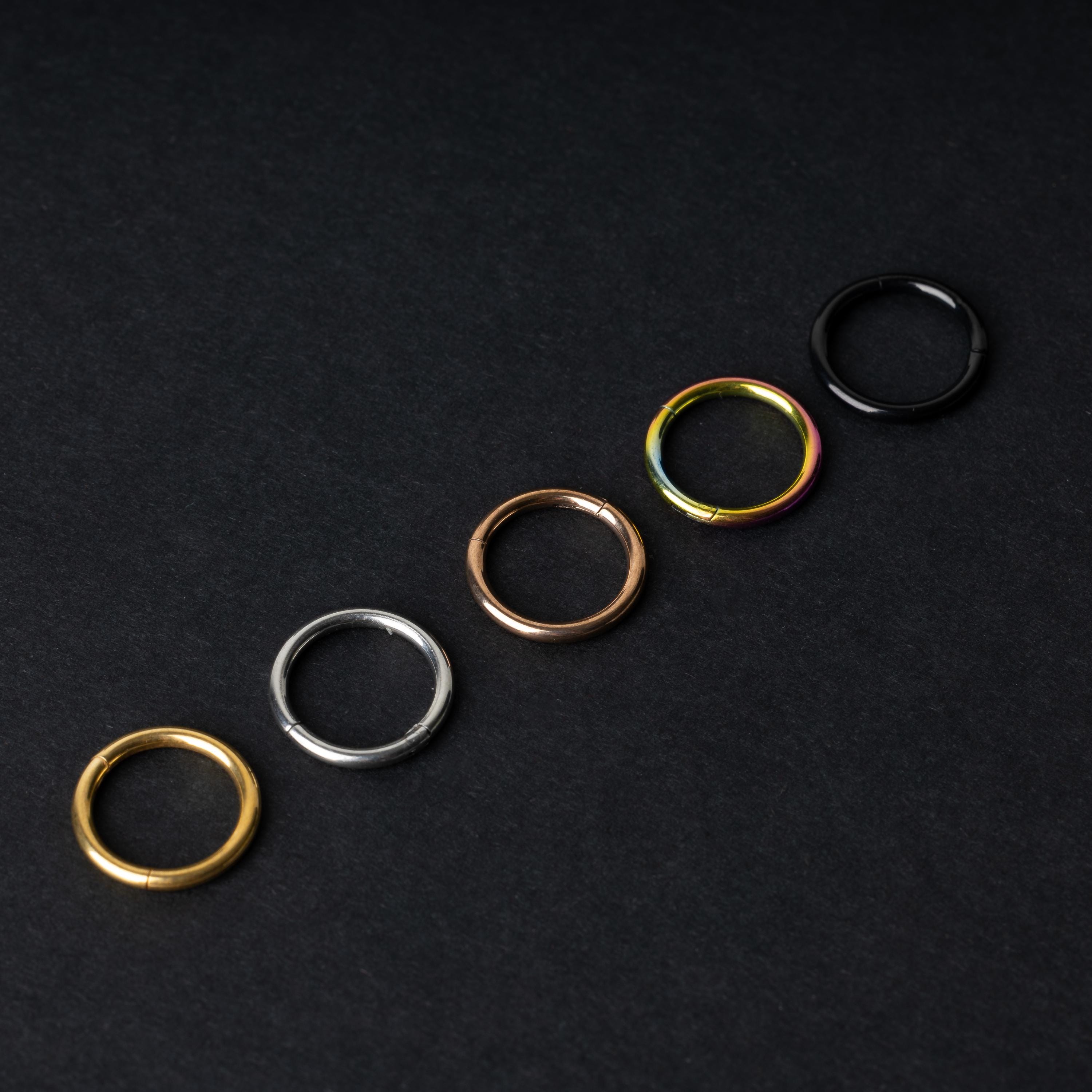 A selection of seamless clicker rings in various finishes – gold, silver, rose gold, rainbow, and black – displayed in a line on a dark background.