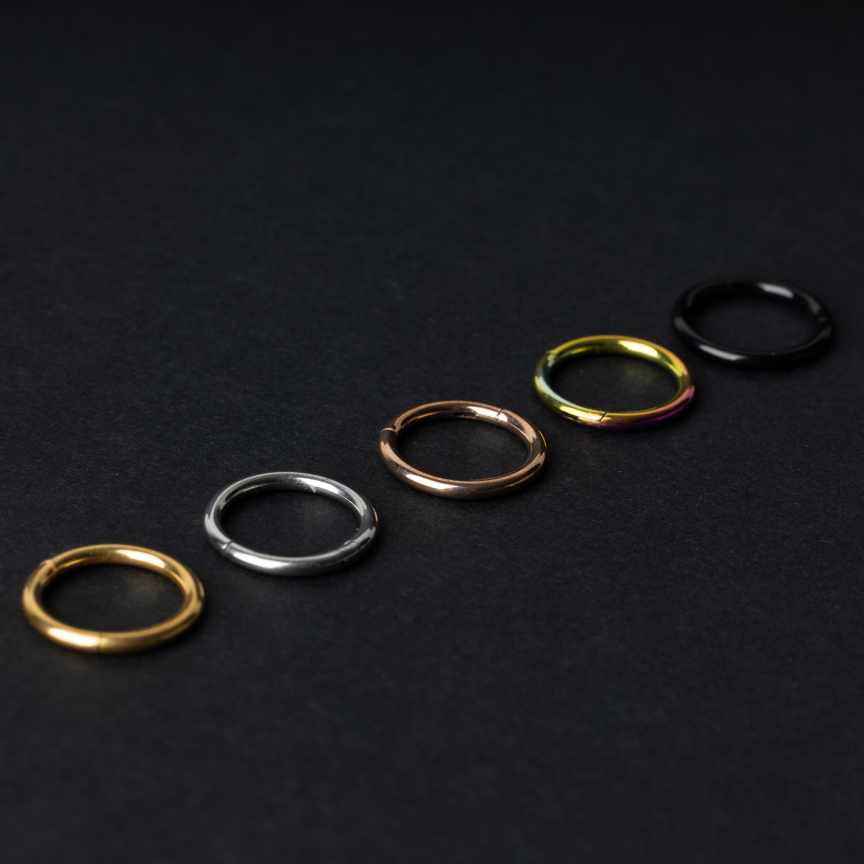 A selection of seamless clicker rings in various finishes – gold, silver, rose gold, rainbow, and black – displayed in a line on a dark background.