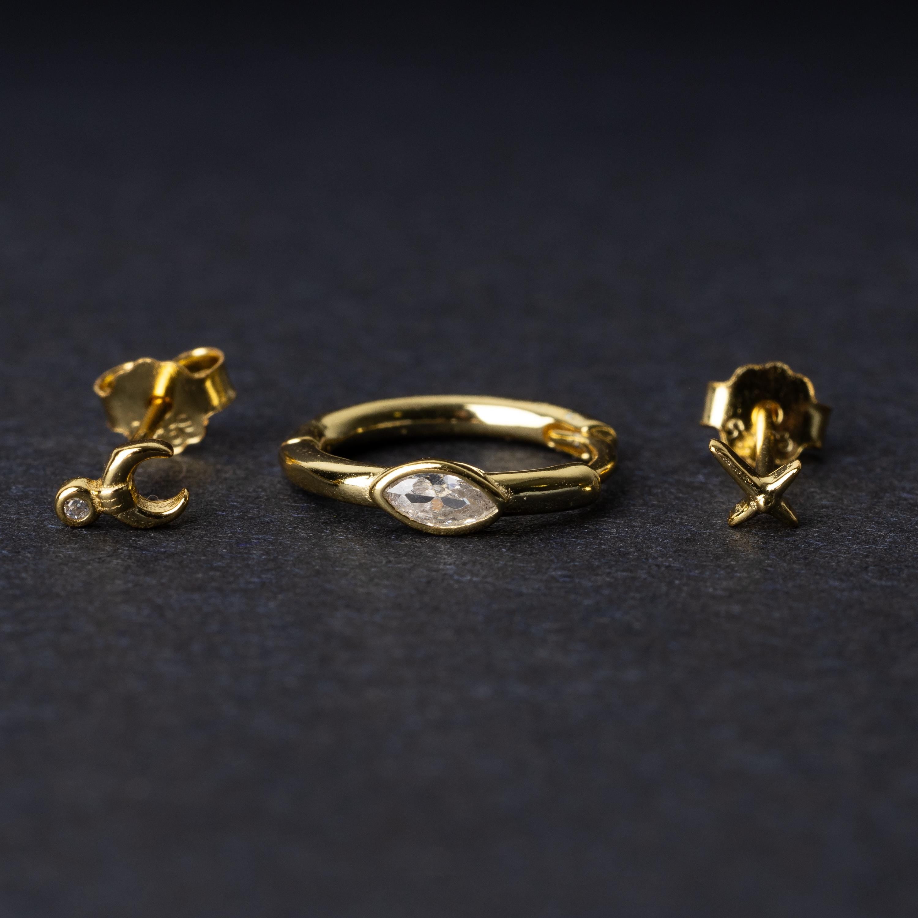 A gold-tone earring set displayed on a dark matte background, featuring a hoop with a marquise-cut crystal, a crescent moon stud with a crystal accent, and a minimalist cross stud.