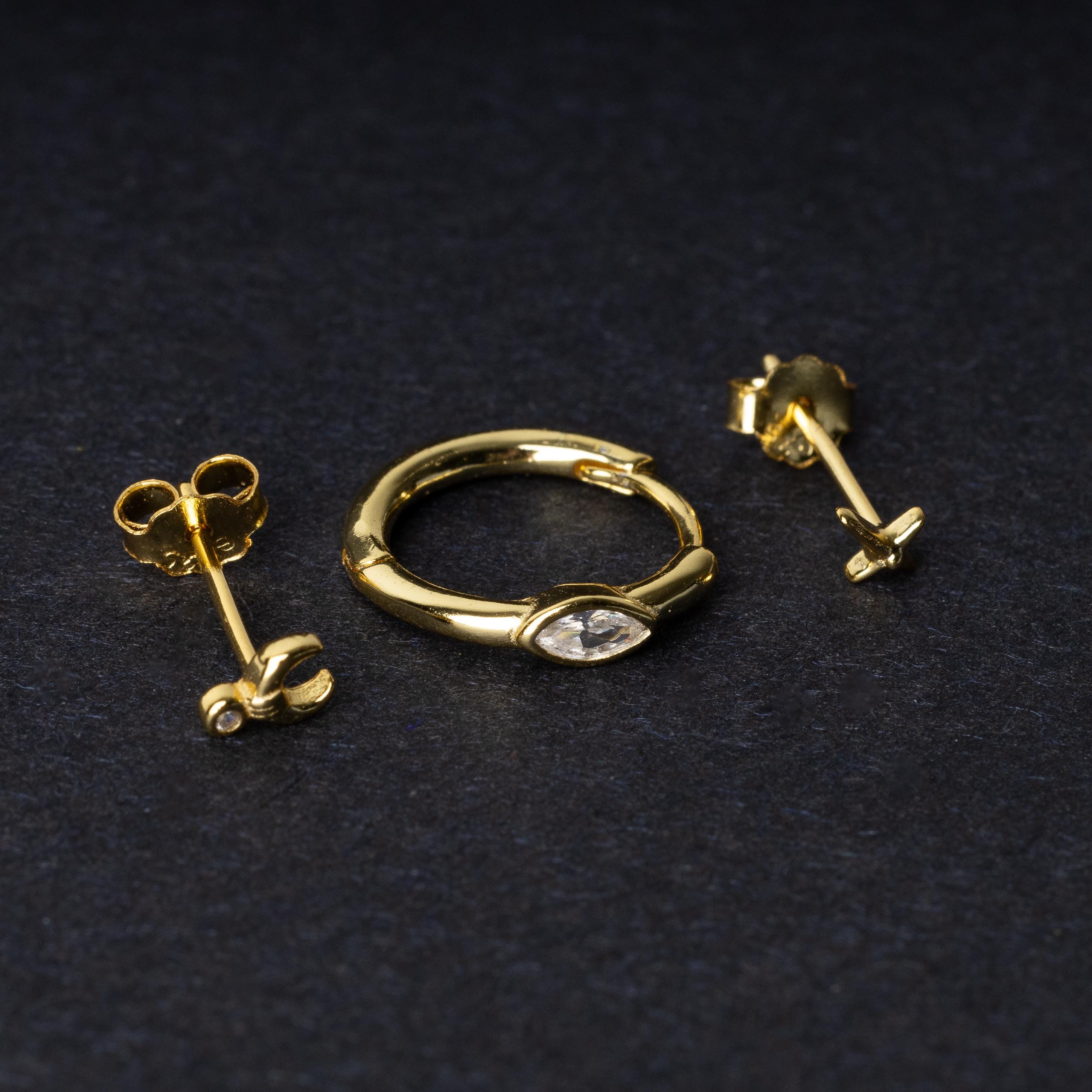 A gold-tone earring set displayed on a dark matte background, featuring a hoop with a marquise-cut crystal, a crescent moon stud with a crystal accent, and a minimalist cross stud.
