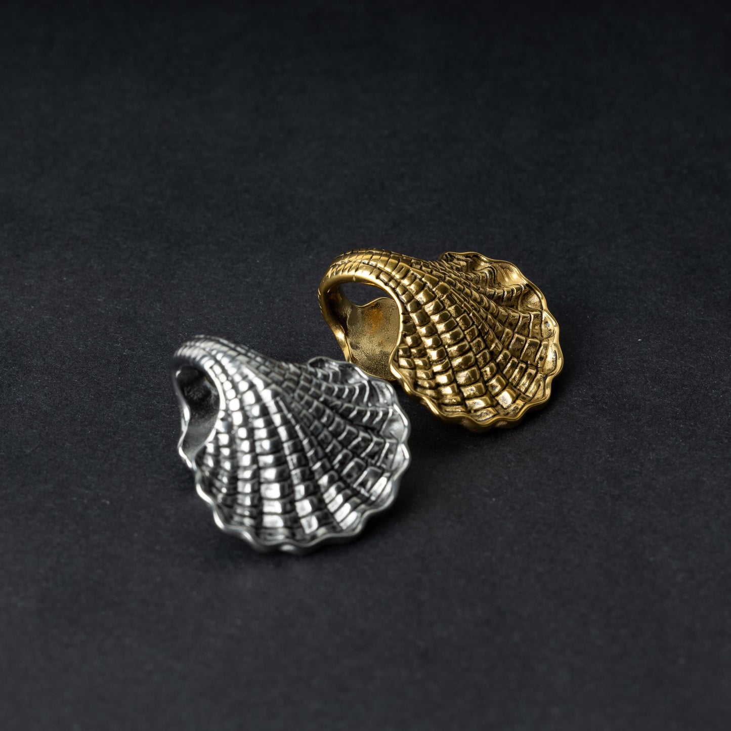 Two seashell-shaped ear weights, one in polished silver and one in polished gold, with detailed textures and a bold design, displayed on a dark background.
