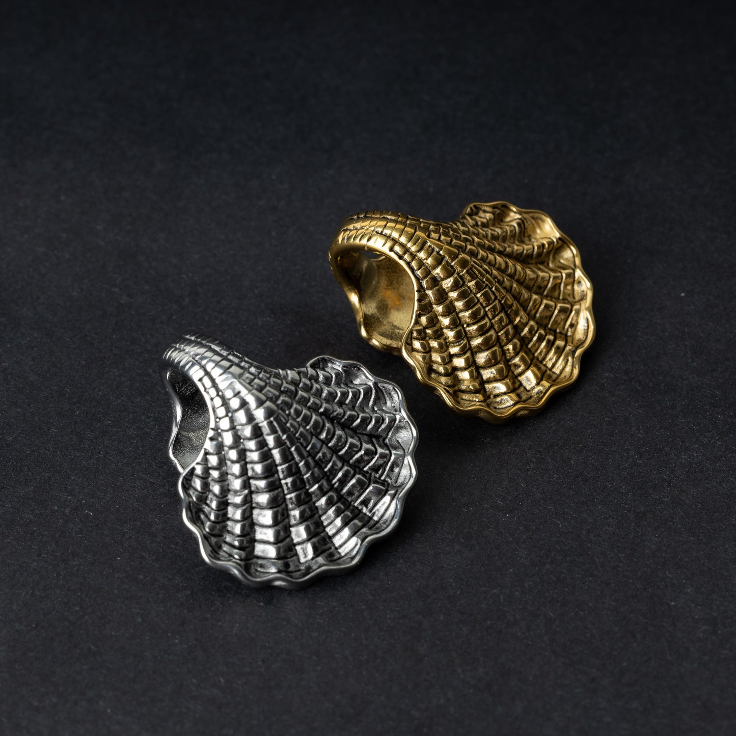 Two seashell-shaped ear weights, one in polished silver and one in polished gold, with detailed textures and a bold design, displayed on a dark background.