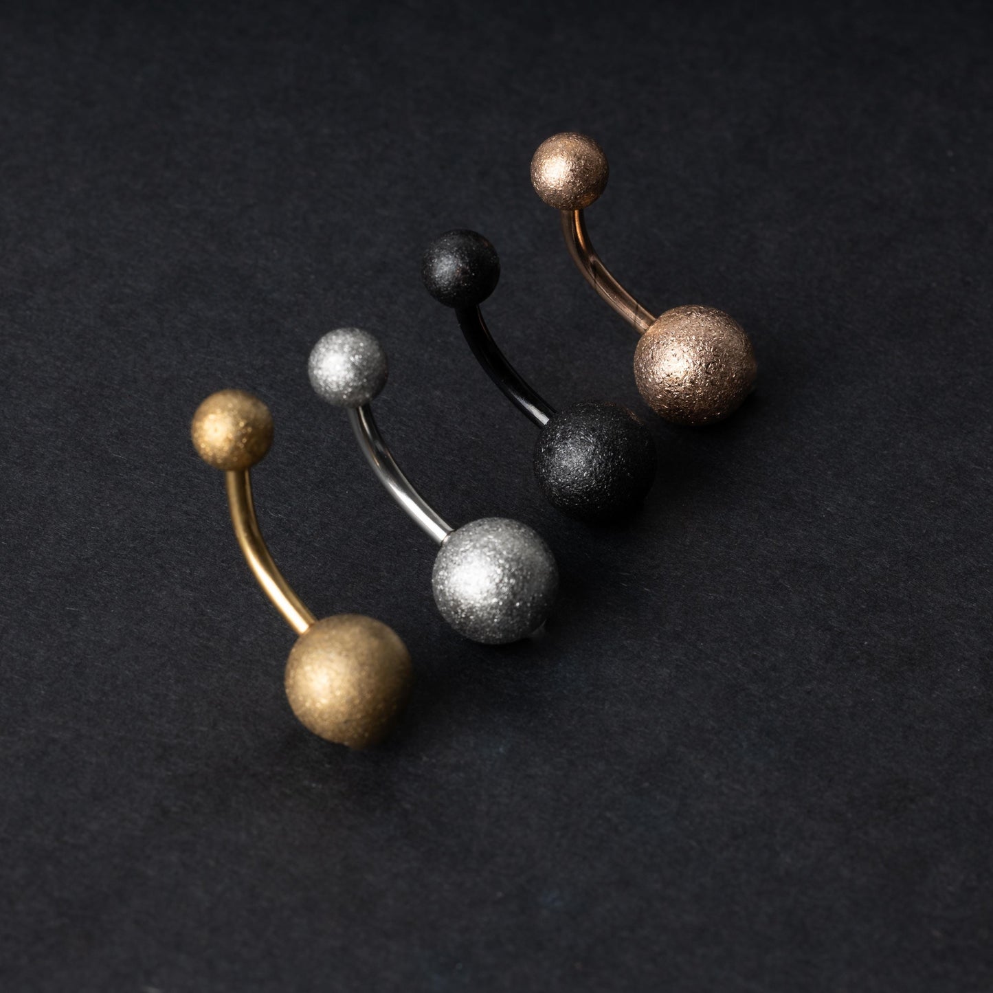 Four curved barbells with shimmering textured ball ends in gold, silver, black, and rose gold finishes, displayed on a dark matte background.