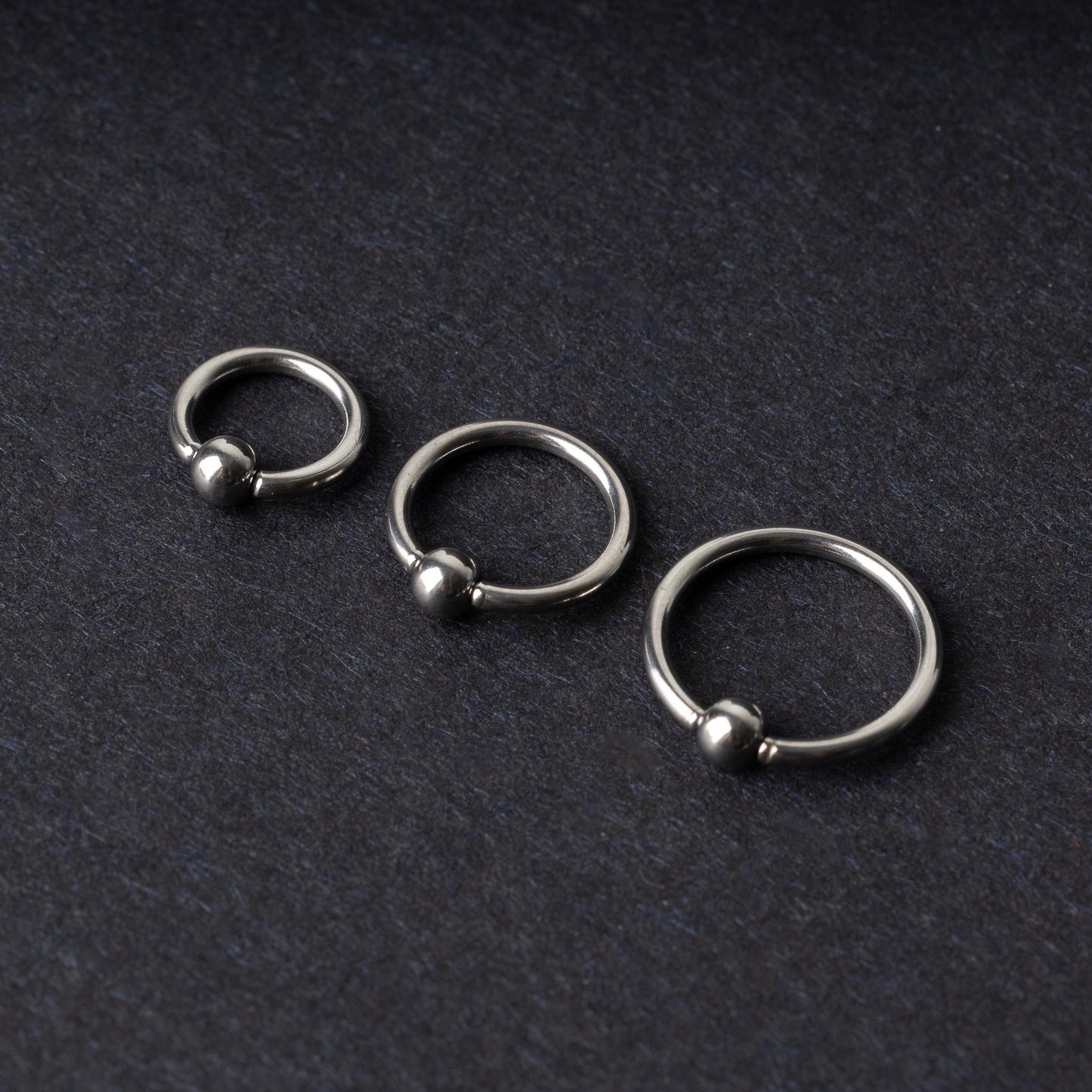 Three polished stainless steel captive bead rings of varying sizes displayed on a dark matte background.