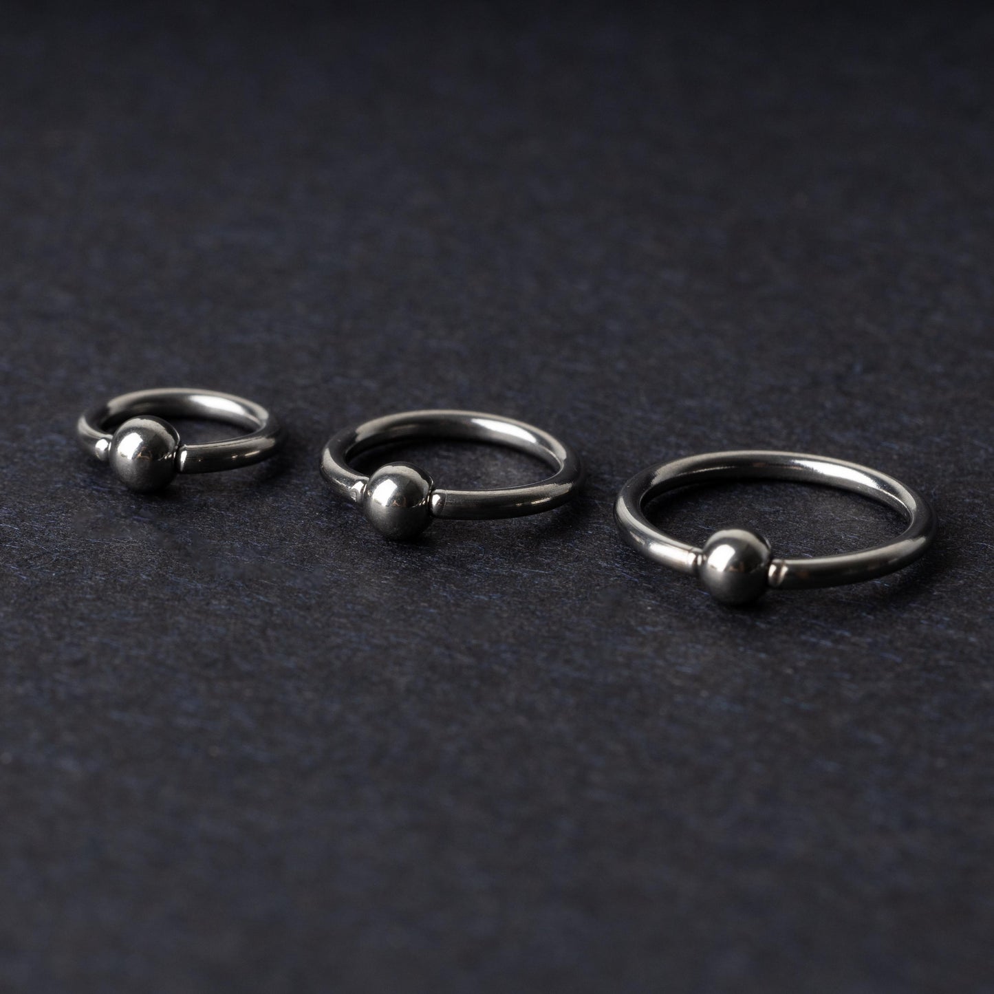 Three polished stainless steel captive bead rings of varying sizes displayed on a dark matte background.