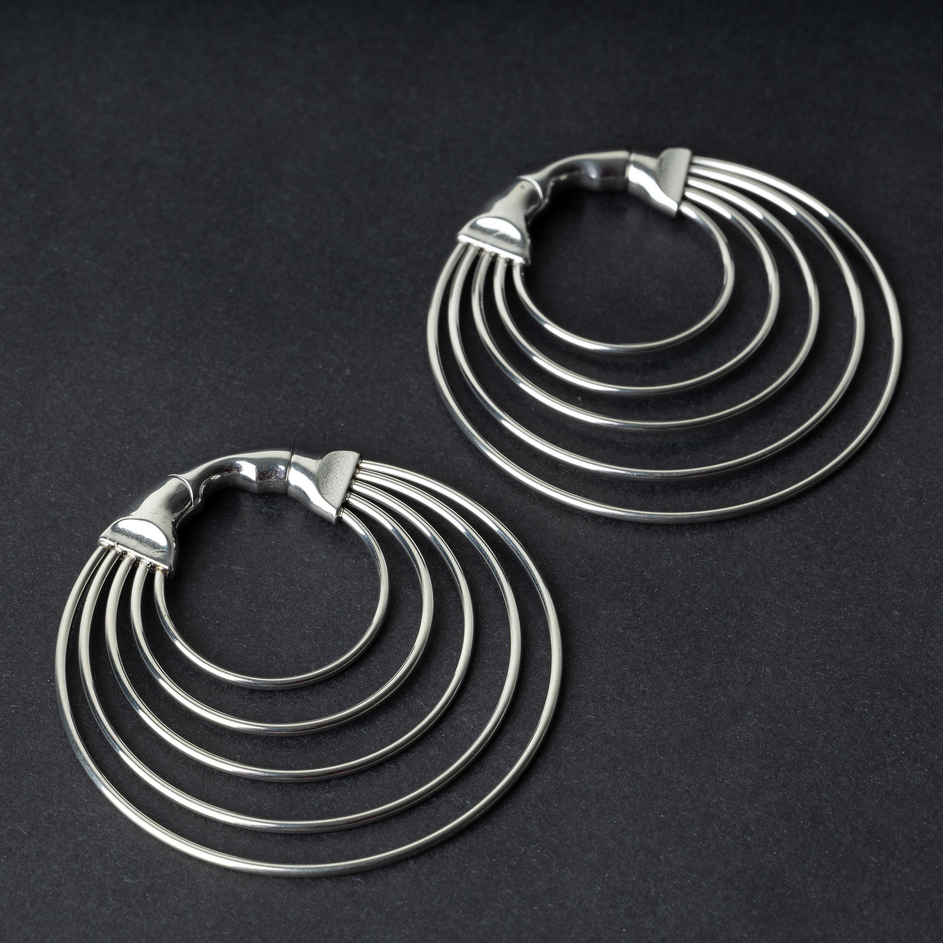 A pair of multi-hoop ear weights in polished silver, featuring five cascading hoops and a seamless hinge closure, displayed on a dark background.