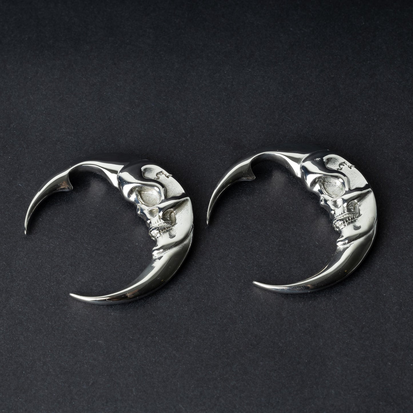 A pair of crescent moon-shaped ear weights in polished silver with detailed moon face designs, displayed on a dark background.