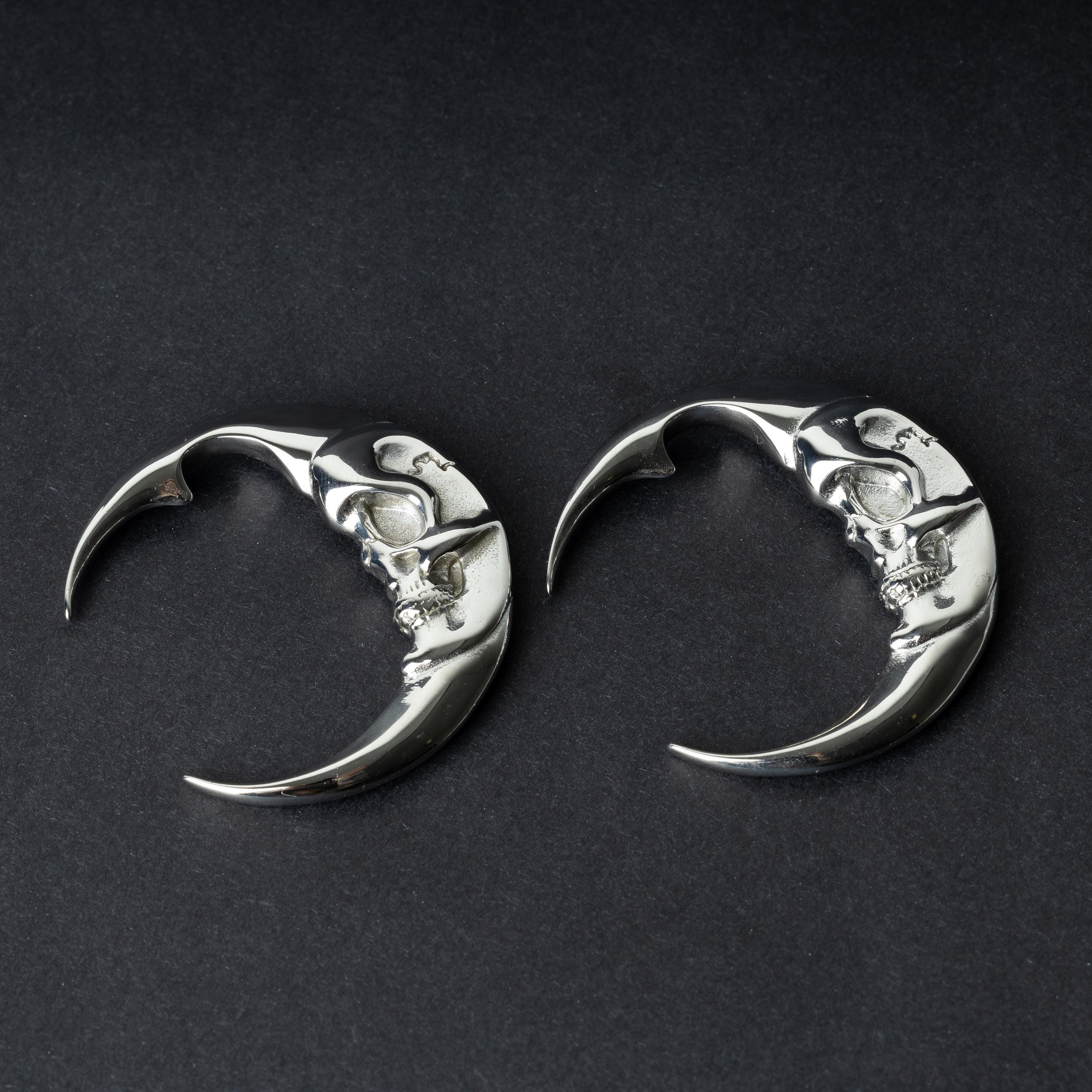 A pair of crescent moon-shaped ear weights in polished silver with detailed moon face designs, displayed on a dark background.