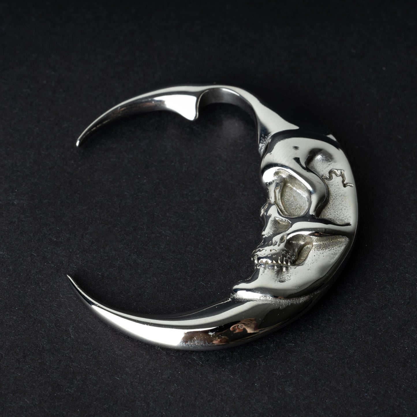 A pair of crescent moon-shaped ear weights in polished silver with detailed moon face designs, displayed on a dark background.