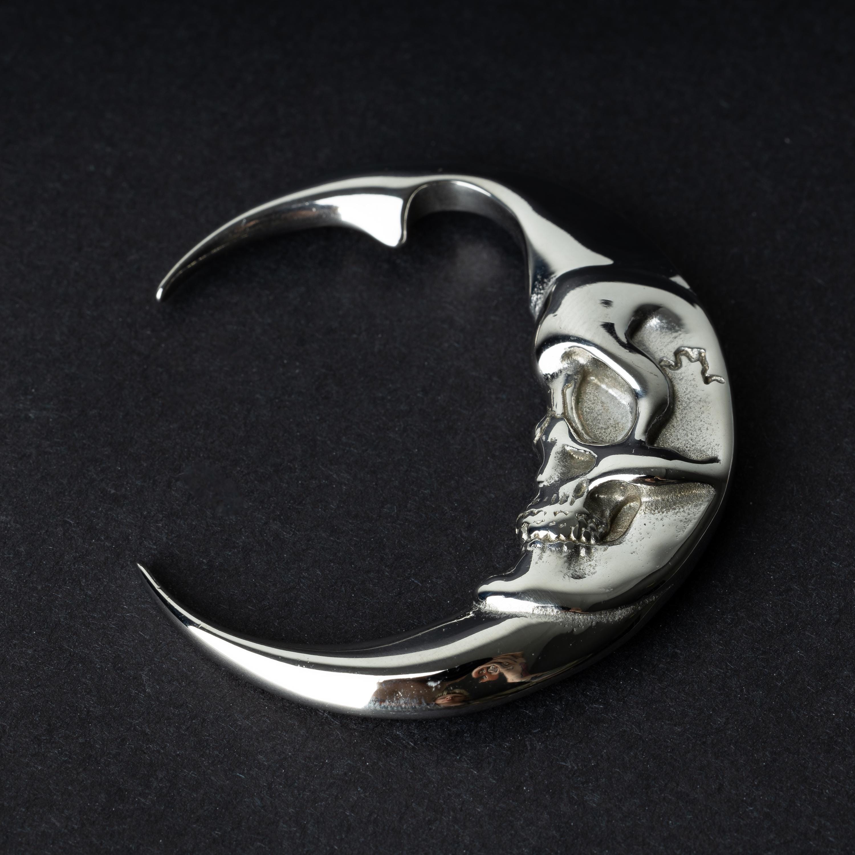 A pair of crescent moon-shaped ear weights in polished silver with detailed moon face designs, displayed on a dark background.